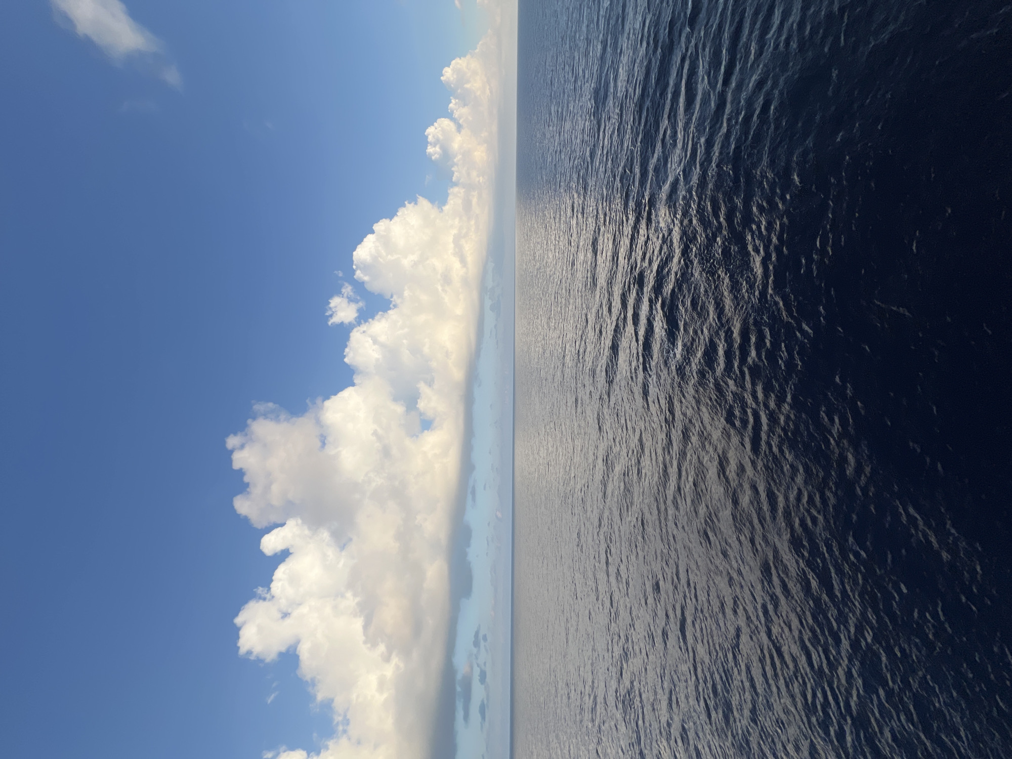 The image depicts a vast expanse of the ocean under a bright blue sky dotted with fluffy white clouds. The horizon line is clearly visible, separating the deep blue of the sea from the lighter blue of the sky. The water has a textured surface with gentle ripples, reflecting the sunlight. The clouds are large and voluminous, with some appearing brighter and more illuminated than others, suggesting a sunny day. The scene evokes a sense of openness and tranquility.