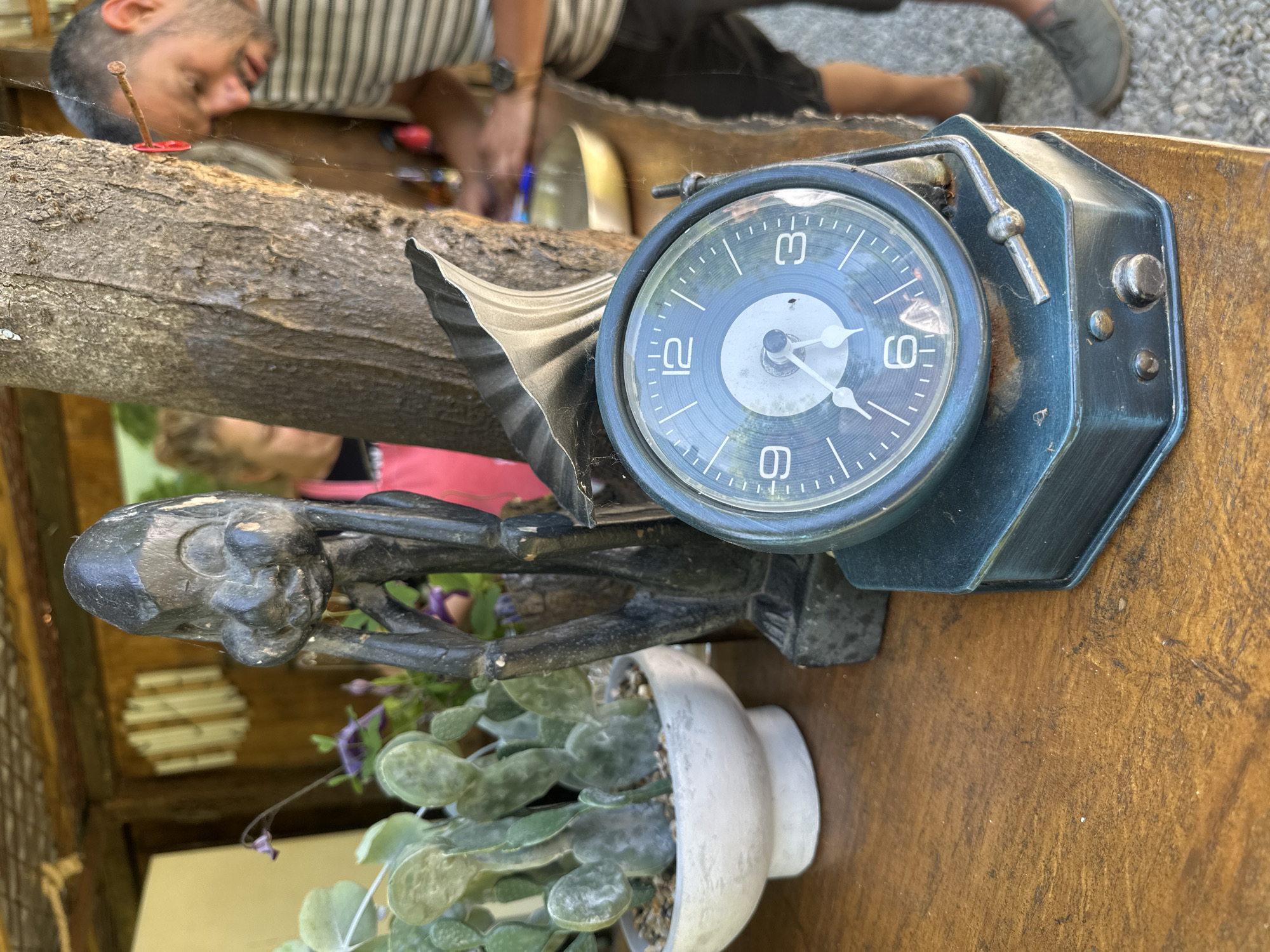 An unconventional, rustic display outdoors. In the foreground is a vintage, blue clock face with white hands and numbers (3, 6, 9, 12) and a matching metal housing. It sits atop a dark statuette of a skeleton-like figure, which sits above a white pot containing a small succulent plant. A rough tree branch is nearby. In the background, a man in a striped shirt and dark shorts lies down on a surface, seemingly working on something, but is slightly out of focus. There is another individual in a pink top partially visible above the skeleton-like figure.
While the location within Podgorica, Montenegro, is not explicitly identified in the image, the scene appears to be at an outdoor marketplace or craft stall, perhaps a tourist location given the unique assemblage of objects.