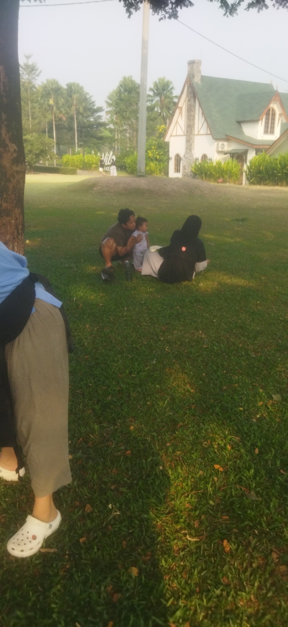 This image depicts a family enjoying a day outdoors in a park setting, possibly at the Martha Tilaar Garden in Depok, Indonesia. In the foreground, a person wearing a blue top and grey pants is partially visible, with their left foot in a white Croc shoe. To their right, a man is squatting and holding a young child, who is wearing a light blue and white patterned outfit. Another person, wearing a black hijab and black top, is seated on the grass next to them. Behind the group, a sign that appears to read "Lakes" is visible amidst lush greenery and palm trees. In the background, a charming white house with a green roof and stone accents is prominent, suggesting it's part of a resort or themed area within the park. The scene is set on a well-maintained lawn, bathed in soft, natural light.