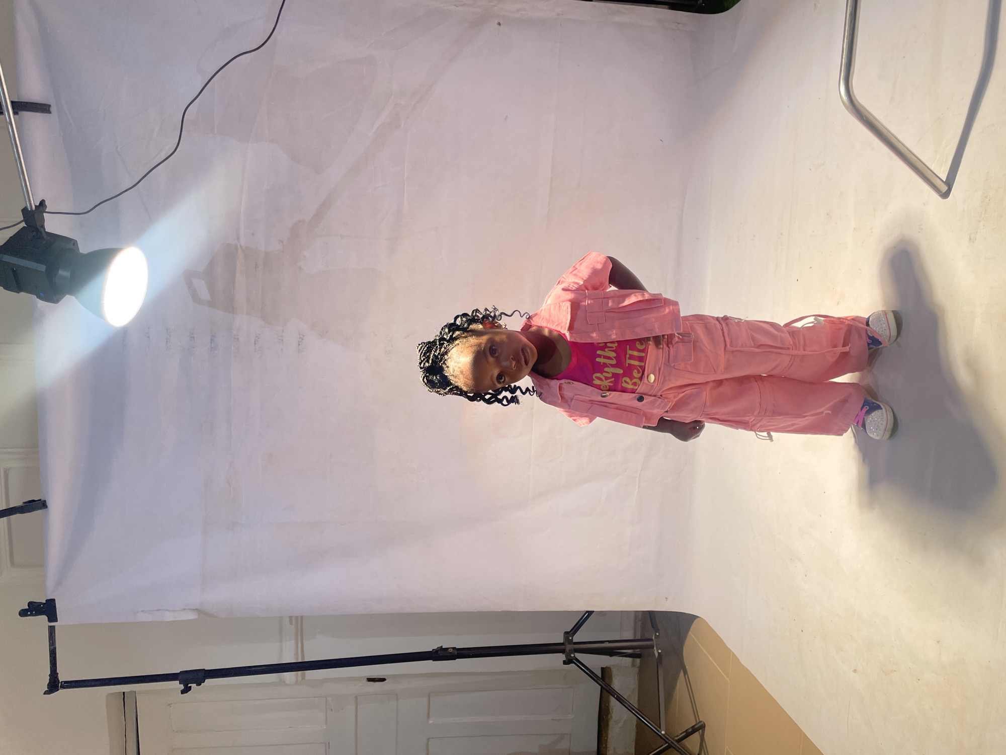 A full-length shot shows a young Black girl in a pink ensemble, posed in what appears to be a photography studio. She is smiling and has braids. A backdrop is visible, as well as studio lighting equipment. The location in Jos, Nigeria is inferred based on the absence of contextual cues in the photo. The setting is a studio with lighting gear. No other people are visible, the main subject is the young girl posing in front of the studio backdrop.