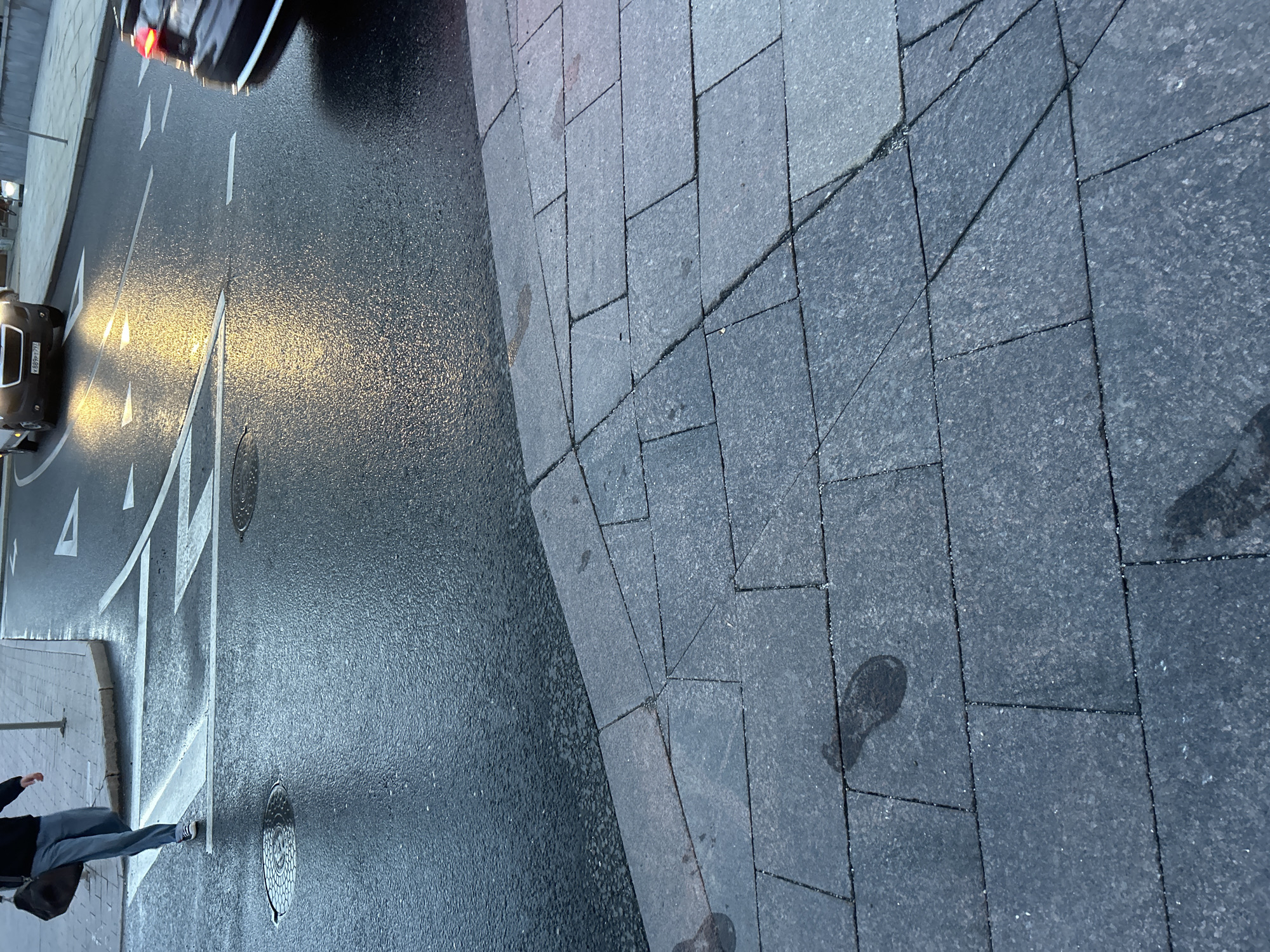 The image shows a street scene in Unknown, Unknown, with a view from a slightly elevated perspective looking down at a wet road and adjacent sidewalk. The road is asphalt, marked with white lane lines and directional arrows. Car headlights reflect brightly off the wet surface. To the right of the road is a sidewalk paved with gray rectangular stones. The sidewalk appears to be wet, with dark patches suggesting dampness. A person is partially visible in the lower left corner of the frame, walking along the road. A dark-colored car is visible on the left side of the road, with its headlights illuminating the wet surface. Another vehicle is partially visible in the upper left corner of the frame. The overall scene suggests an urban environment, possibly after a rain.