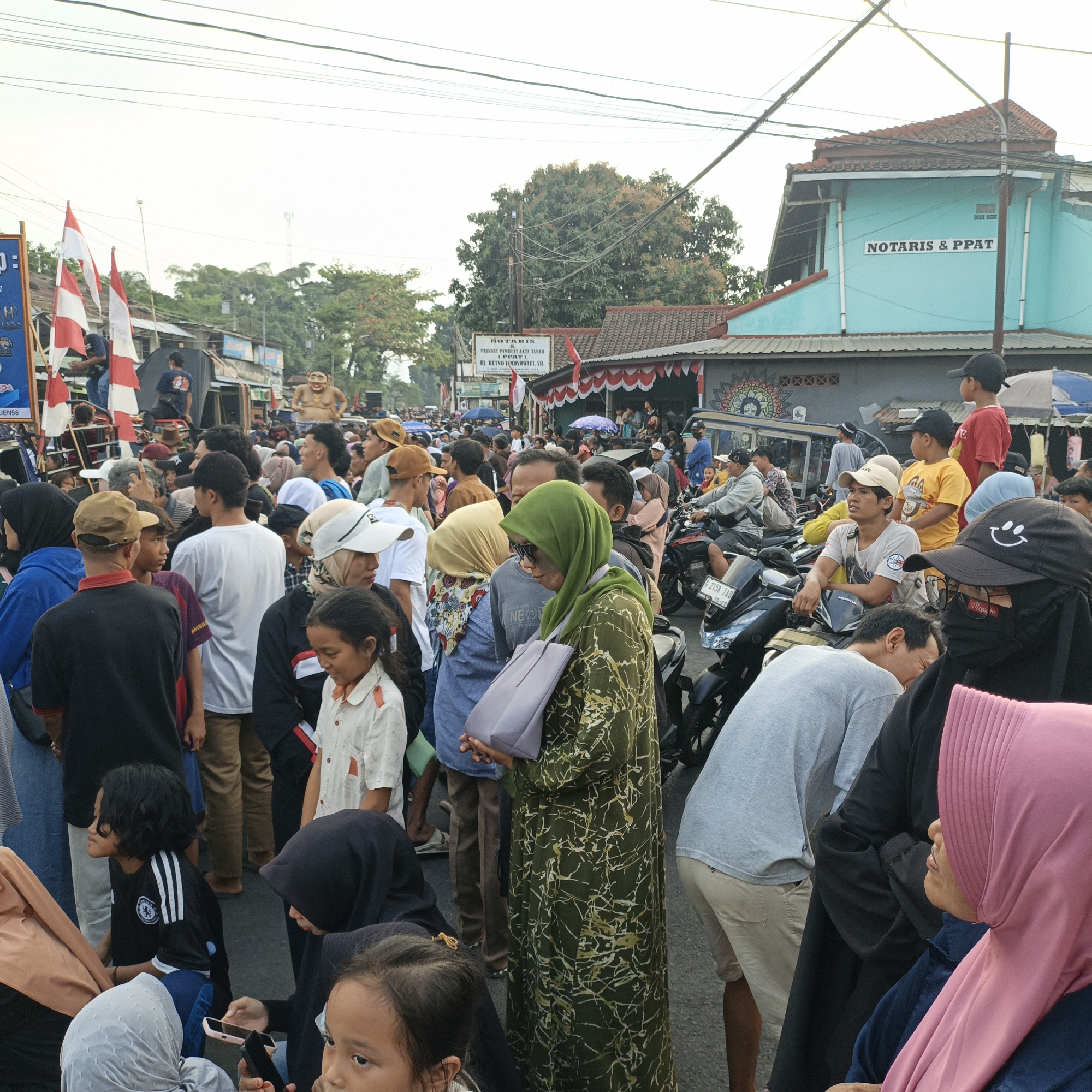 The image depicts a crowded street scene in Randudongkal, Indonesia. The foreground is occupied by a diverse group of people, predominantly dressed in casual clothing, including several wearing hijabs. A young girl in a black and white striped soccer jersey is visible in the lower left quadrant. To her right, a woman in a black hijab holds a mobile phone. In the middle ground, a large gathering of men and women are observing something to their left, indicated by their turned heads and focused gazes. Several individuals are wearing hats, and one person is wearing a distinctive green patterned dress. In the background, a building with "NOTARIS & PPAT" signage is visible, along with Indonesian flags. Motorcycles are parked and some are in motion, suggesting a busy urban environment. The overall atmosphere appears to be one of a public event or gathering, possibly a parade or festival, given the flags and the celebratory mood suggested by the large number of people present.