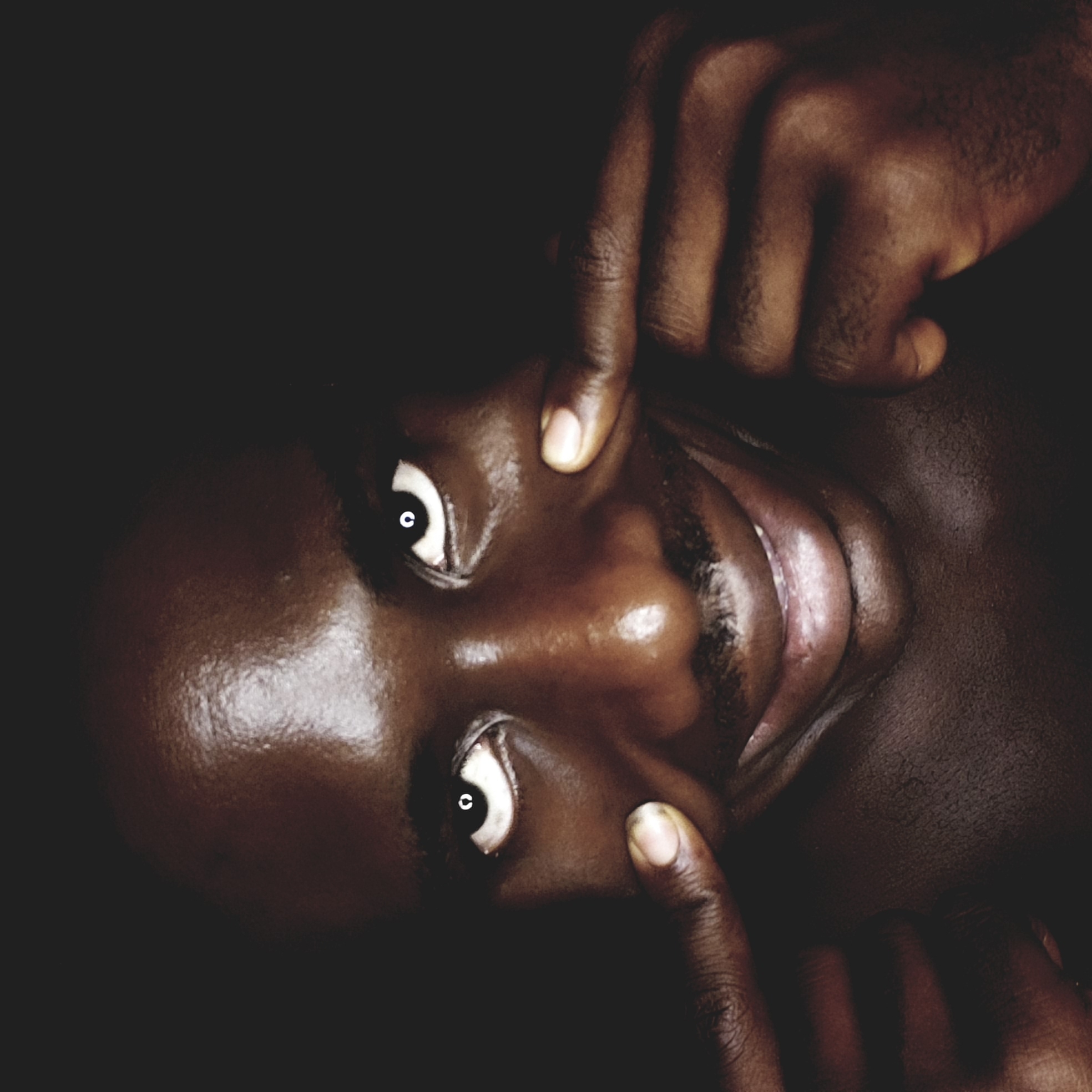 An extreme close-up photograph features a dark-skinned person lying on their back, their face illuminated against a stark black background. The person's eyes are wide open, and their fingers are positioned on their cheeks, pulling them slightly outward. The skin appears oily, reflecting light. The person has a slight moustache and the mouth is open. No specific details indicate that the location is Narayi, Nigeria; this cannot be confirmed.