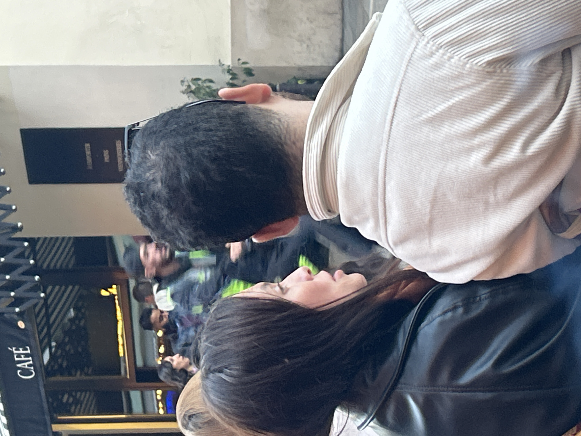 A medium shot depicts two individuals standing close together outdoors, with a partially visible "CAFÉ" sign suggesting a public or commercial area. The woman, dressed in a black leather jacket and with dark hair, looks upwards. The man, wearing a cream-colored shirt, is positioned to the left, and a crowd of people can be seen behind them near the entrance of the café.