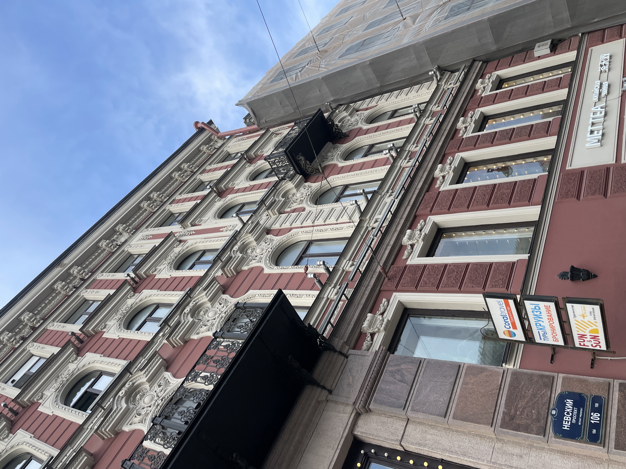 The image shows a building facade in Saint Petersburg, Russia. The architecture is ornate, featuring a red and white color scheme, arched windows, decorative moldings, and wrought-iron balconies. Scaffolding covers the upper portion of the building. Several signs are attached to the building, including "Coraltravel," "Fun & Sun," and a sign written in Cyrillic script that translates to "Chinese Tea." Street signs indicate that the building is located on Nevsky Prospekt, at street numbers 104, 106, and 108. The sky is partly cloudy.