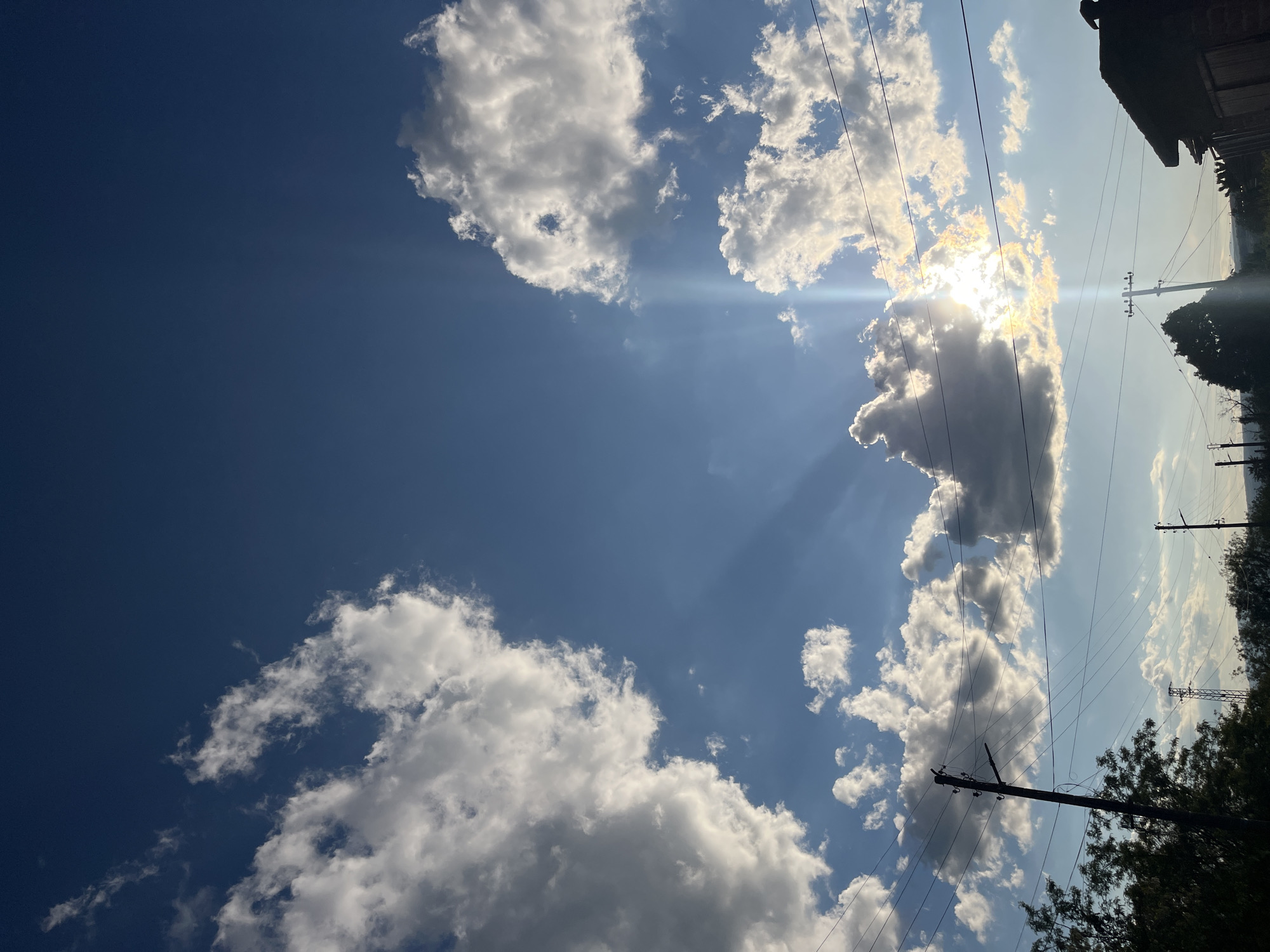 The image captures a partly cloudy sky above Lozarevo, Bulgaria. The sun is partially obscured by a large cloud formation. Overhead power lines cross the sky. On the right side of the image, foliage and portions of structures, including an old building, are visible. The sky appears blue.