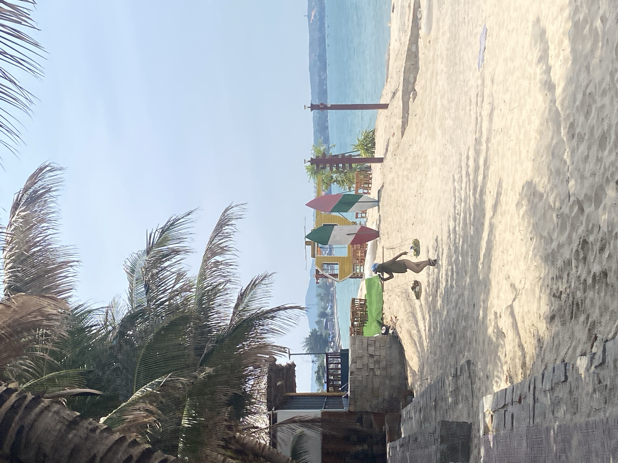An individual is walking across a sandy beach in Phu Thuy Ward, Vietnam, carrying two baskets suspended from a pole across their shoulders. The person is wearing a dark top, shorts, and a light-colored head covering. The beach is wide with light-colored sand, and palm trees with fronds overhang the scene from the left, casting shadows. In the background, the turquoise ocean stretches towards a hazy horizon with a distant landmass. Several structures are visible on the beach, including a thatched-roof building on the left, and a yellow and red booth-like structure further back. Decorative surfboards stand upright near the booth, and small tables and chairs are set up on a patch of green astroturf. The sky is a pale blue, clear and bright, suggesting a sunny day.