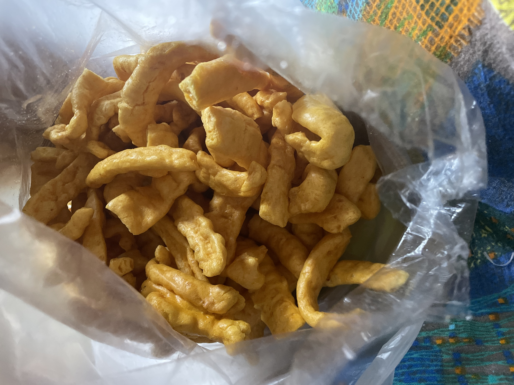 An overhead, close-up, eye-level, natural light, full shot shows a plastic bag filled with a pile of yellow, puffy, extruded snacks. The snacks are irregular in shape, with some appearing twisted or curved. The plastic bag is mostly transparent, allowing a clear view of the snacks inside. The bottom of the bag rests on a colorful, patterned surface, possibly a tablecloth or placemat, with blues, greens, oranges, and yellow hues. The scene is indoors, with no discernible features indicating a specific location within Davao City, Philippines, or any other geographical context. There are no people or animals present in the image, and no actions are being performed other than the snacks being contained within the bag. The overall impression is of a casual, everyday snack item.