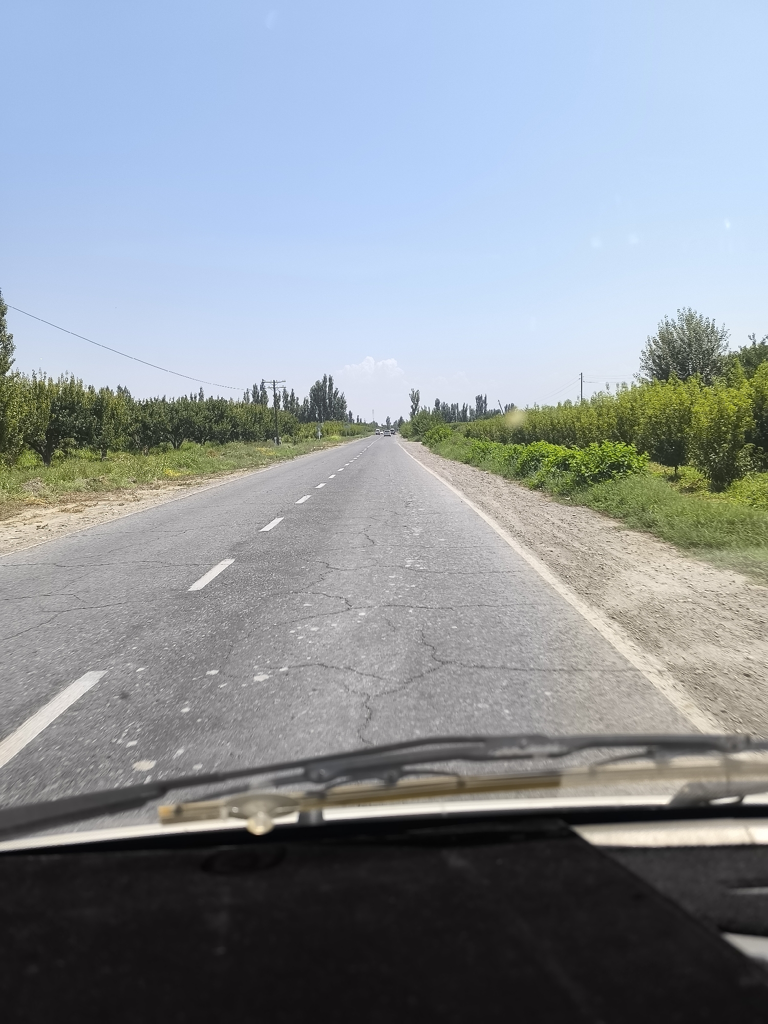 The image shows a view from inside a vehicle looking out at a paved road in Hudayarhan, Uzbekistan. The road has white dashed lines indicating lanes. To the left of the road, there is a grassy verge and then rows of trees. To the right of the road, there is also a grassy verge and dense green vegetation, with more trees further in the distance. The sky is clear and blue with a few faint clouds. There are a couple of distant vehicles on the road. The foreground of the image is dominated by the dashboard and windshield wipers of the vehicle.