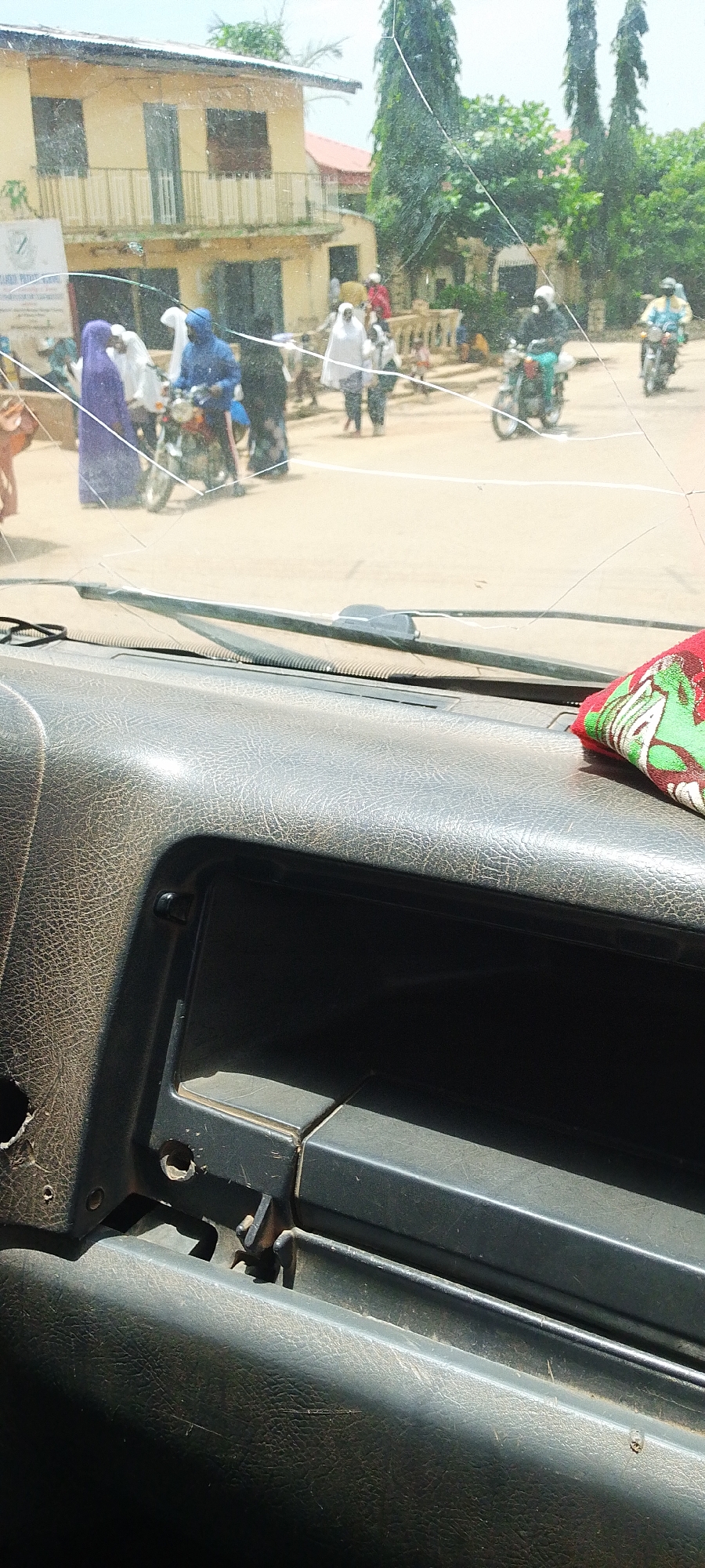 This image depicts a street scene in Kwang, Nigeria, viewed from inside a vehicle through a cracked windshield. Several individuals are visible on the street, some walking and others on motorcycles. A building with a pale yellow facade and balconies is present in the background. The street appears to be unpaved, with a sandy or dusty surface. The overall impression is of a bustling local environment.