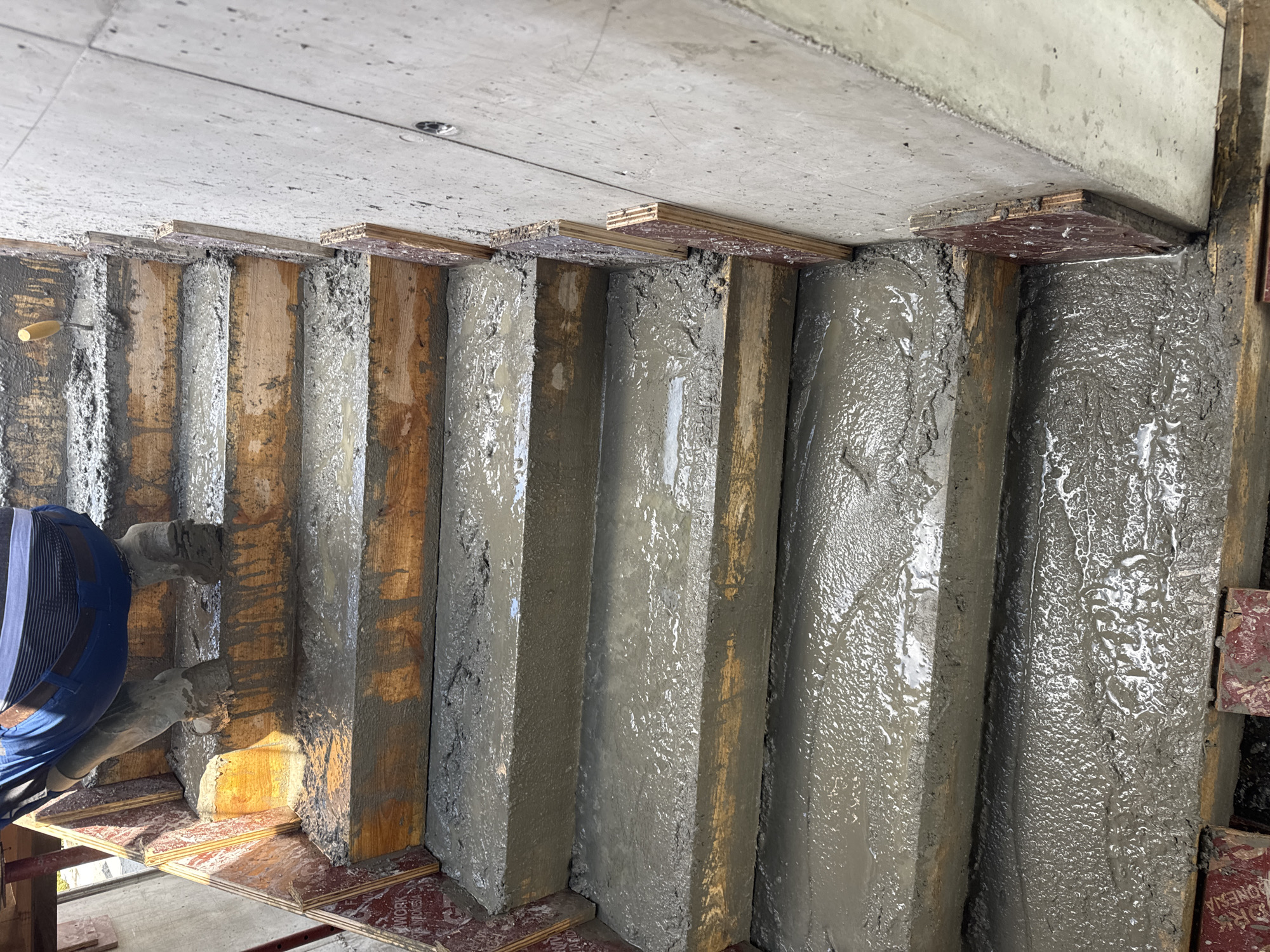 This image captures a construction scene focused on concrete pouring and formwork. The main subjects are fresh, wet concrete contained within vertical wooden forms, and a partially visible worker.
The scene shows several parallel vertical wooden structures, which are acting as formwork for concrete columns or walls. These forms are filled with gray, freshly poured concrete, which appears wet and somewhat glistening, with visible textures and liquid patterns on its surface. Some areas of the concrete appear more settled and smooth, while others retain a rougher finish.
Above the vertical forms, a finished concrete ceiling or slab is visible, showing a mottled gray texture. Small, thin plywood pieces are strategically placed between the top edges of the wooden forms and the overhead concrete, likely serving as shims or part of the formwork bracing system.
On the left side of the image, the lower body and arms of a worker are visible, clad in blue work trousers and a blue top. The worker is holding what appears to be a tool, possibly a concrete vibrator or a hose, suggesting they are actively involved in consolidating the concrete or managing its flow. A small trowel with a wooden handle is resting on the concrete near the worker.
Beneath the vertical forms, a base of horizontal plywood sheets is visible, supporting the formwork. Some of these sheets are stained a reddish-brown color. Text is partially visible on one of these plywood pieces, showing fragments like "ORK" and "ONESIA," which could indicate "FORM WORK INDONESIA" or similar product branding.
The image depicts a daytime setting within a construction environment, consistent with ongoing building activity. The overall impression is one of manual labor and the precise handling of materials during the concrete casting phase of construction in Plovdiv, Bulgaria.