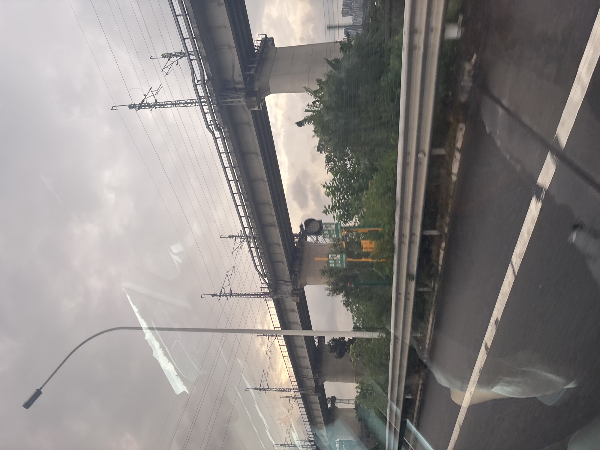 The image depicts an elevated railway track supported by concrete pillars, stretching across a cloudy sky in Baiyun District, China. The railway is adorned with overhead electrical lines and transmission towers. Below the track, a dense growth of green trees and foliage lines the area. A metal guardrail is visible in the foreground, separating the roadway from the embankment. The road surface appears wet, with white lane markings indicating a road. Reflections of the interior of a vehicle are visible on the surface of the image, obscuring parts of the scene. The overall atmosphere is overcast, with hints of sunlight breaking through the clouds, casting a soft glow on the horizon. In the distance, a cluster of high-rise buildings can be seen.