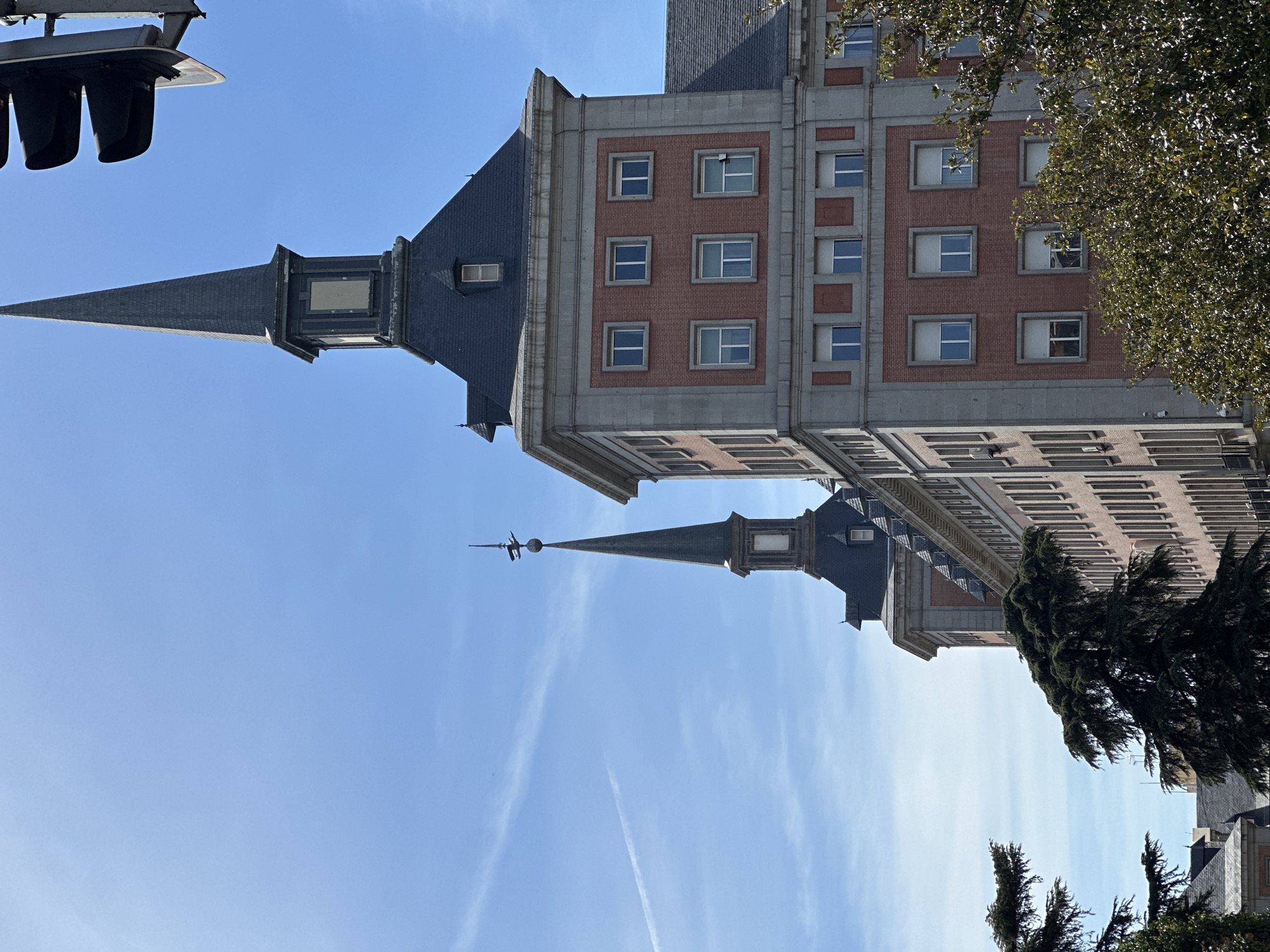 The image shows the Torrespaña building in Madrid, Spain, viewed from below at street level. The building has a distinctive spire and red-brick and grey stone construction. The sky is blue. Parts of trees and a traffic light are visible at the edges of the frame.