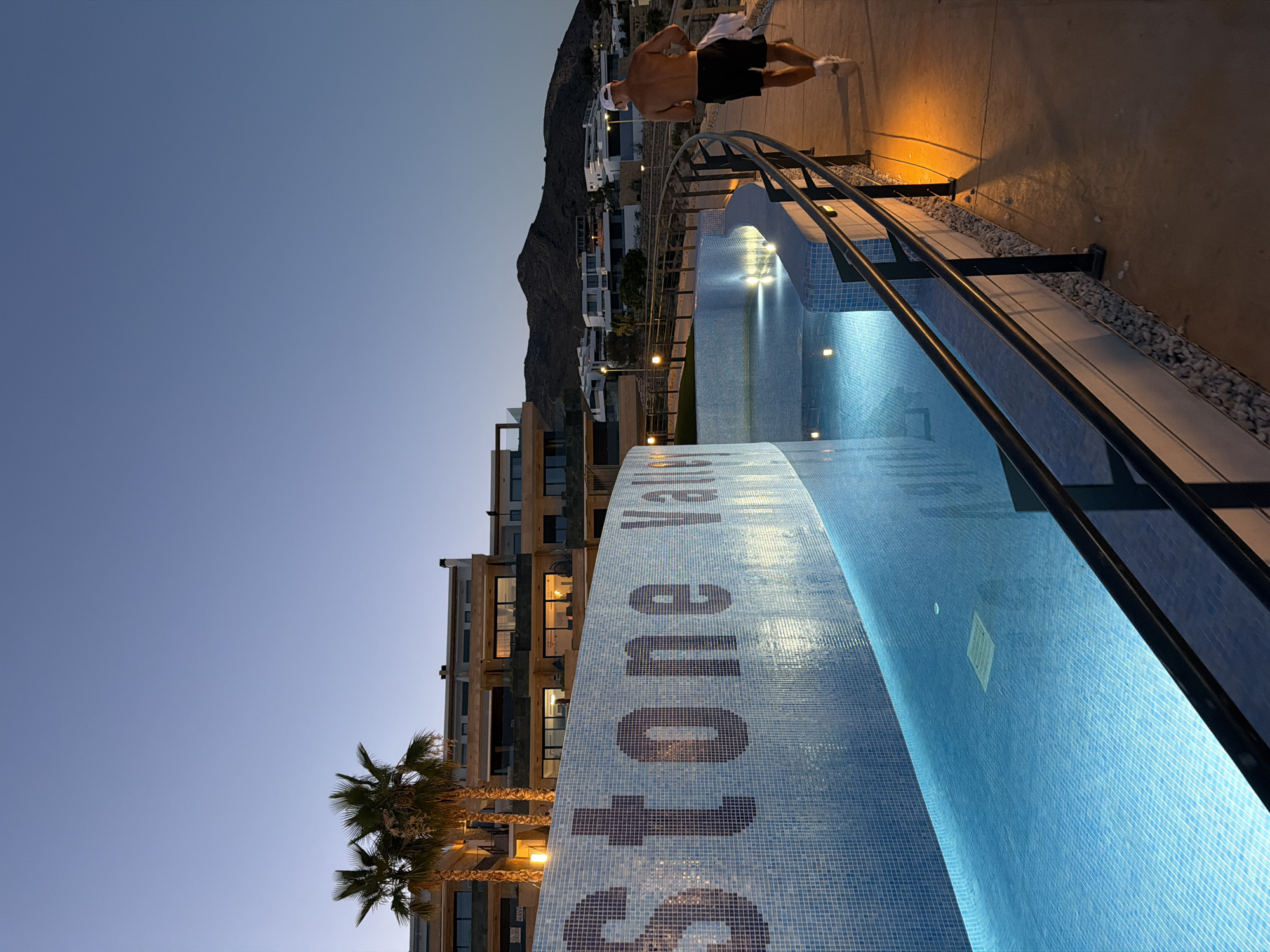 The image shows an outdoor scene at dusk in Finestrat, Spain, likely on the grounds of the "Stone Valley" resort. The foreground features a curved swimming pool with bright blue, illuminated water, tiled with small, light blue mosaic squares. A dark railing runs along the edge of the walkway next to the pool. A man, shirtless with dark shorts and a white cap, is walking away from the camera along this walkway, holding a white towel over his shoulder. Behind the pool, a curved wall is decorated with dark mosaic tiles that spell out "Stone Vall" (likely "Stone Valley"). In the mid-ground, palm trees and modern buildings with illuminated windows are visible. The background displays a hilly, mountainous landscape with more buildings scattered across the slopes, under a clear, deep blue twilight sky. Streetlights with warm yellow glows are visible along the walkway and in the distance, illuminating the scene.