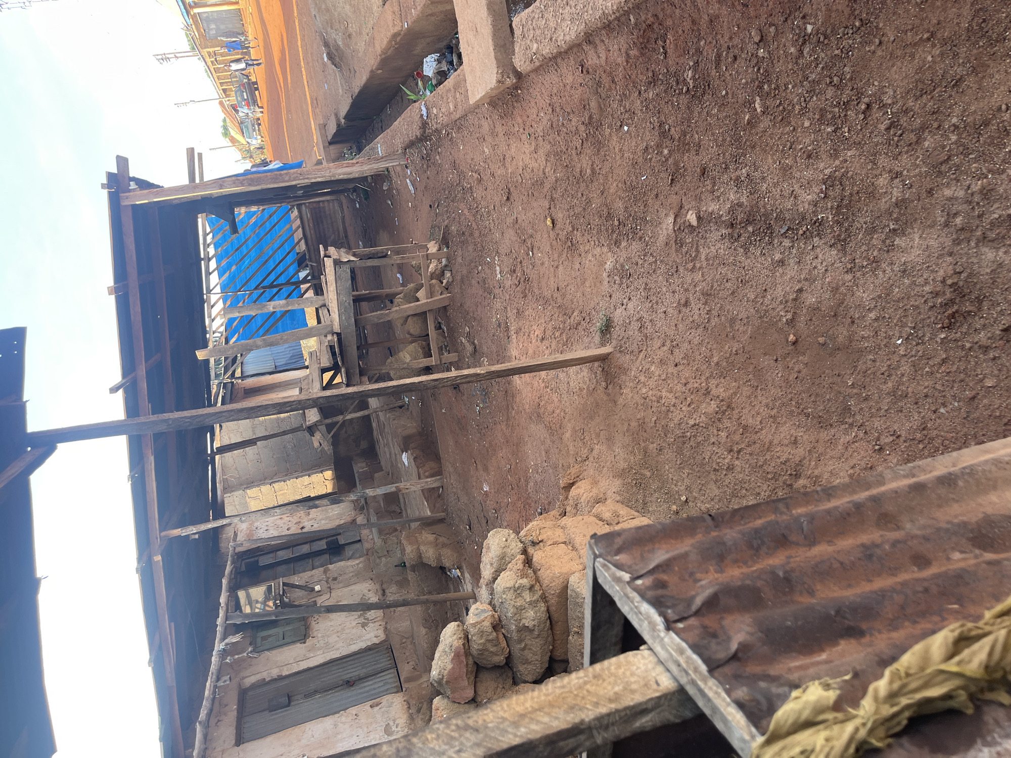This media file captures an unpaved, reddish-brown earthen area in a rural or semi-urban setting, likely Kuru Dungus, Nigeria, during bright daylight.
On the left side of the frame, several rudimentary structures are visible, primarily constructed from wooden beams and planks. One of these structures features a bright blue tarp or corrugated plastic sheeting as part of its roof or wall. The walls of some buildings appear to be made of light-colored blocks or mud, with partially closed windows and doors, some covered with corrugated metal sheets. Near the base of these structures, piles of rough, light-colored rocks or dried mud bricks are stacked.
The ground across the scene is unpaved red soil, interspersed with small rocks and scattered debris, including plastic items and general refuse, particularly noticeable in a shallow ditch or drainage area.
In the right foreground, a rusty corrugated metal sheet, possibly part of a makeshift roof or barrier, is prominently featured, with a crumpled yellowish-brown piece of fabric draped over it.
In the distant background, an unpaved road runs horizontally. Parked vehicles, including a dark sedan and a lighter-colored car, are visible along this road. Several indistinct human figures, likely three or four, can be seen standing or walking near these vehicles. Utility poles with overhead wires are also present further in the background.
The sky is bright and clear, indicating fair weather and daytime conditions. No specific activity or event is taking place; the scene appears static, depicting an everyday environment. There is no visible text in the image.