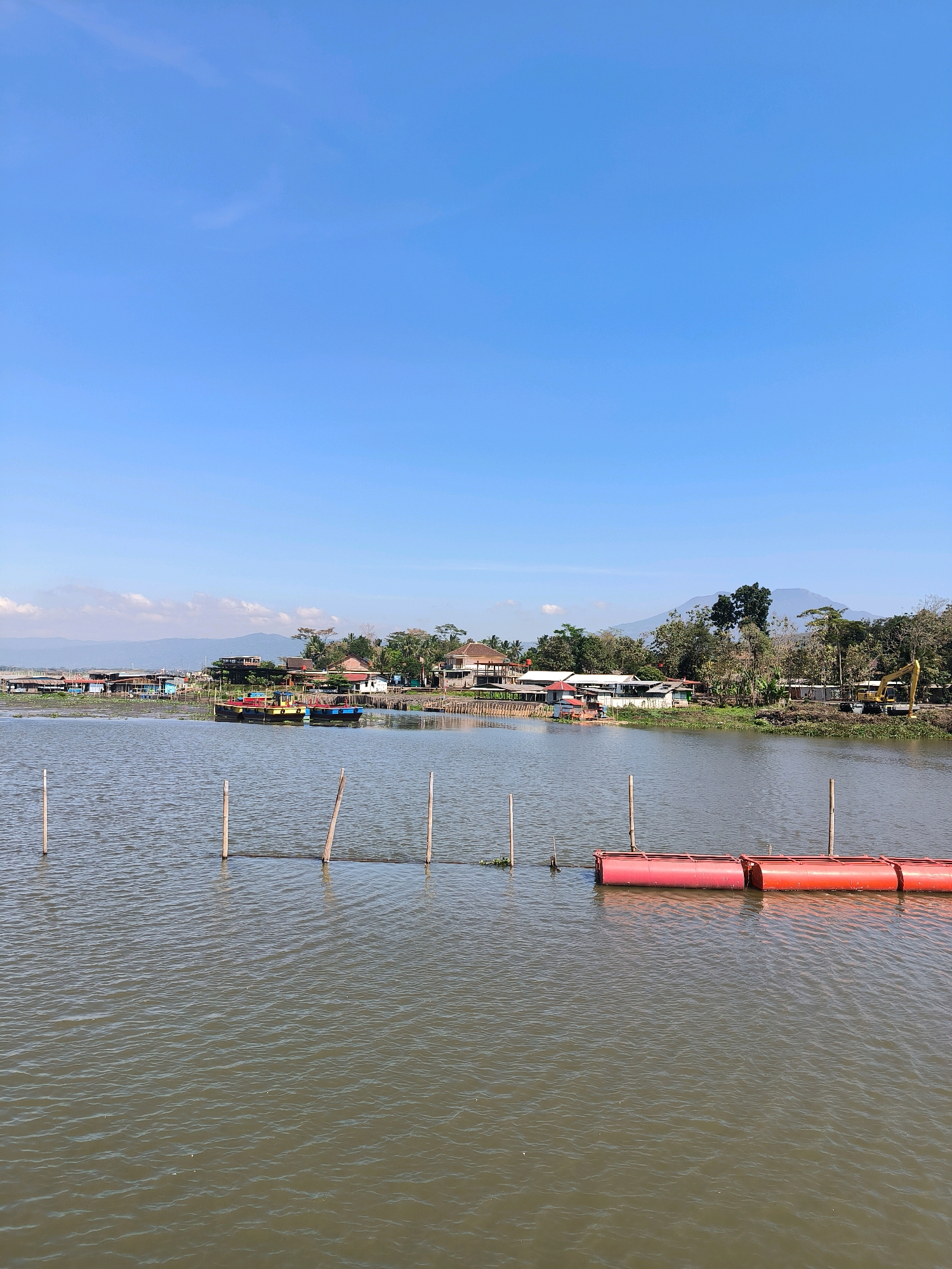 This vertical, eye-level outdoor photograph captures a serene view of the Tuntang River in Tuntang, Indonesia. The image is dominated by a vast expanse of calm, rippling water under a clear, bright blue sky with a few wispy clouds near the horizon. Wooden poles are partially submerged in the water, creating a visual line across the foreground.
In the middle ground, a line of red cylindrical buoys floats on the water. Beyond the buoys, the riverbank is lined with a variety of structures. There are several boats docked, their hulls visible in various colors, including red and yellow. Buildings, appearing to be a mix of houses and businesses, are clustered along the shore, with roofs in shades of red, brown, and white. Trees and vegetation are interspersed among the buildings, with a larger mountain range visible in the distant background. To the right, a yellow excavator is parked on the bank, suggesting ongoing construction or maintenance work. The scene is peaceful and indicative of a riverside community.
