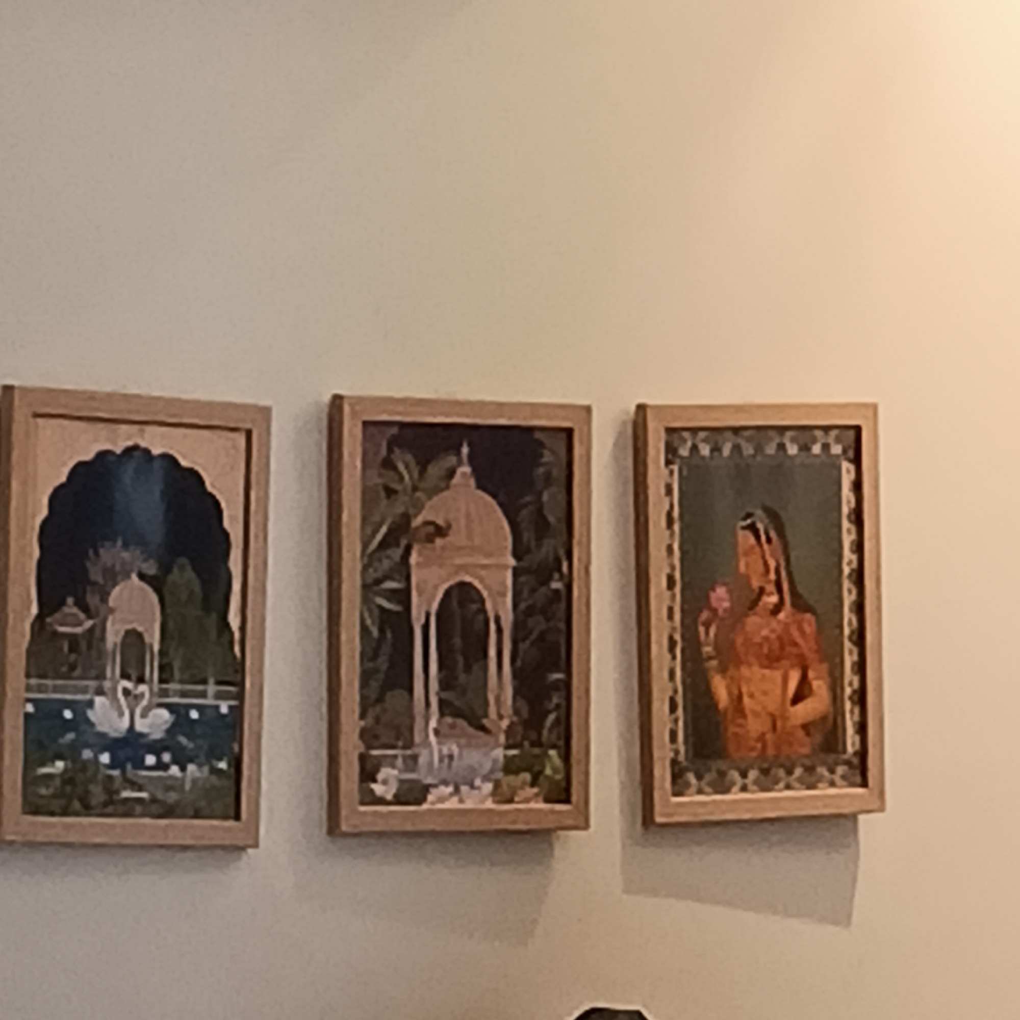 Three framed paintings hang on a light-colored wall. From left to right, the first painting depicts a water scene with two swans and a building under a dark blue arch. The second painting shows a domed structure surrounded by foliage. The third painting features a woman holding a flower. The frames are all similar in style, appearing to be made of light wood. The setting is indoors. No people are visible other than the depiction in the third painting. The location is unknown, the city of Calangute is only mentioned in the instructions.