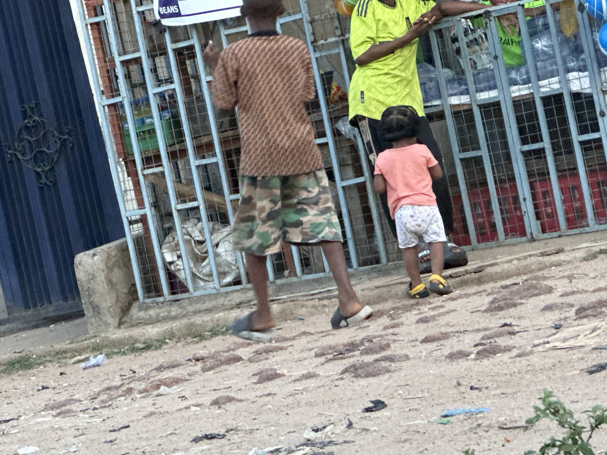 A medium shot captures three people standing in front of a metal-barred structure with a corrugated iron door to the left. The foremost individual is a boy seen from the back, clad in a brown patterned shirt and camouflage shorts, stepping forward in gray and white flip-flops. Next to him, a child in a peach-colored shirt and patterned shorts is also facing away from the camera, wearing yellow and black shoes. Adjacent to the child, a figure in a bright yellow shirt stands partly behind the metal barrier, with only the torso visible. The ground is a light brown color, strewn with debris and small patches of greenery. A sign bearing the word "BEANS" is visible above the figures behind the metal bars. The location appears to be an urban setting in Unknown, Unknown.
