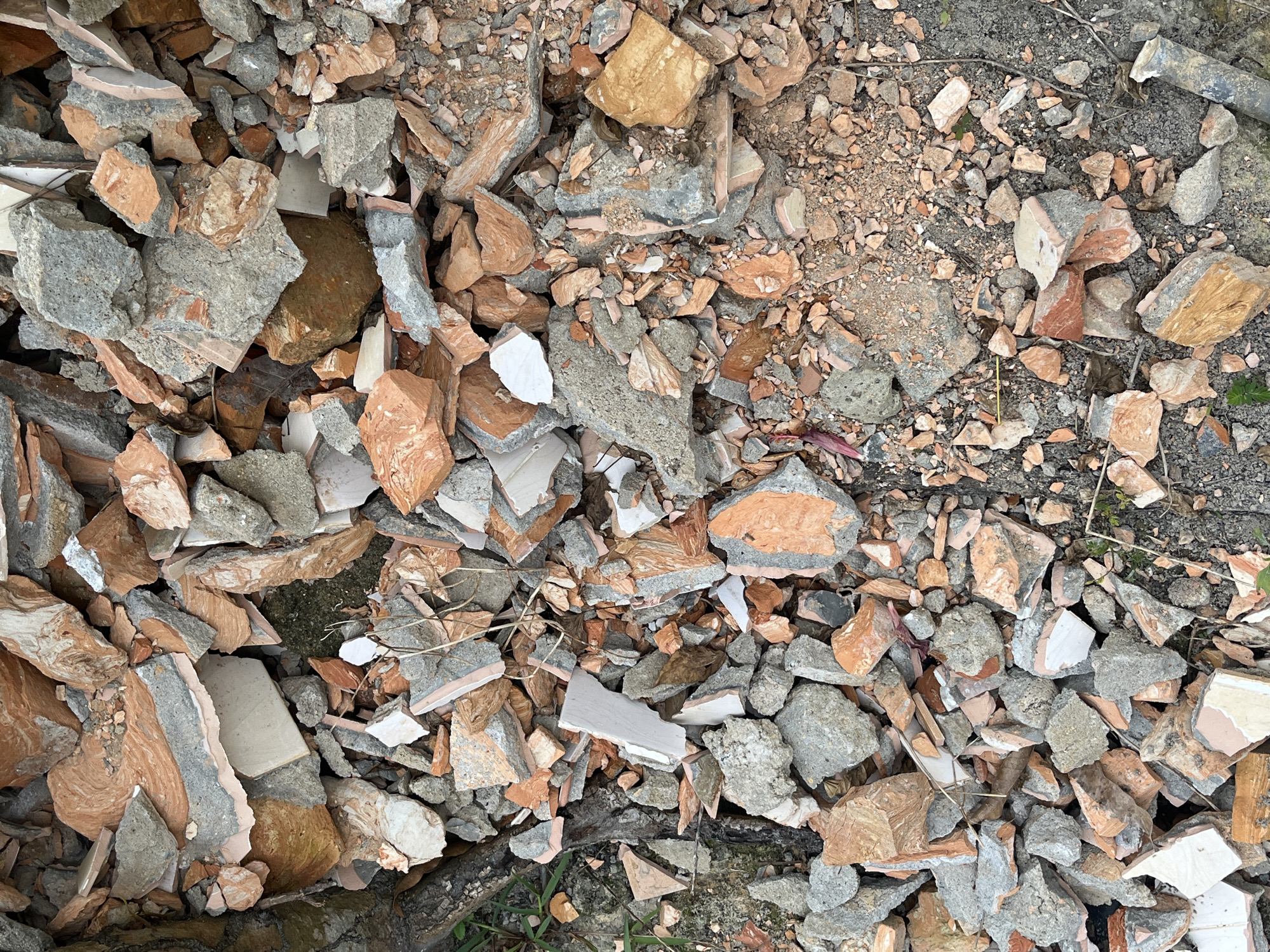 The image is a top-down, full-frame shot of a pile of rubble, likely from a demolition or construction site. The debris consists of broken pieces of concrete, brick, and what appears to be ceramic tile. The colors are predominantly grey, various shades of orange and red from the bricks and tiles, with some white and light brown pieces. The rubble is scattered across a rough, granular surface, possibly dirt or gravel, with some dry vegetation and a dark branch on the left side. The composition is densely packed with fragments of varying sizes, creating a textured and chaotic visual. The image does not contain any people or indicate a specific location beyond being a general construction or demolition debris pile. The setting appears to be outdoors.