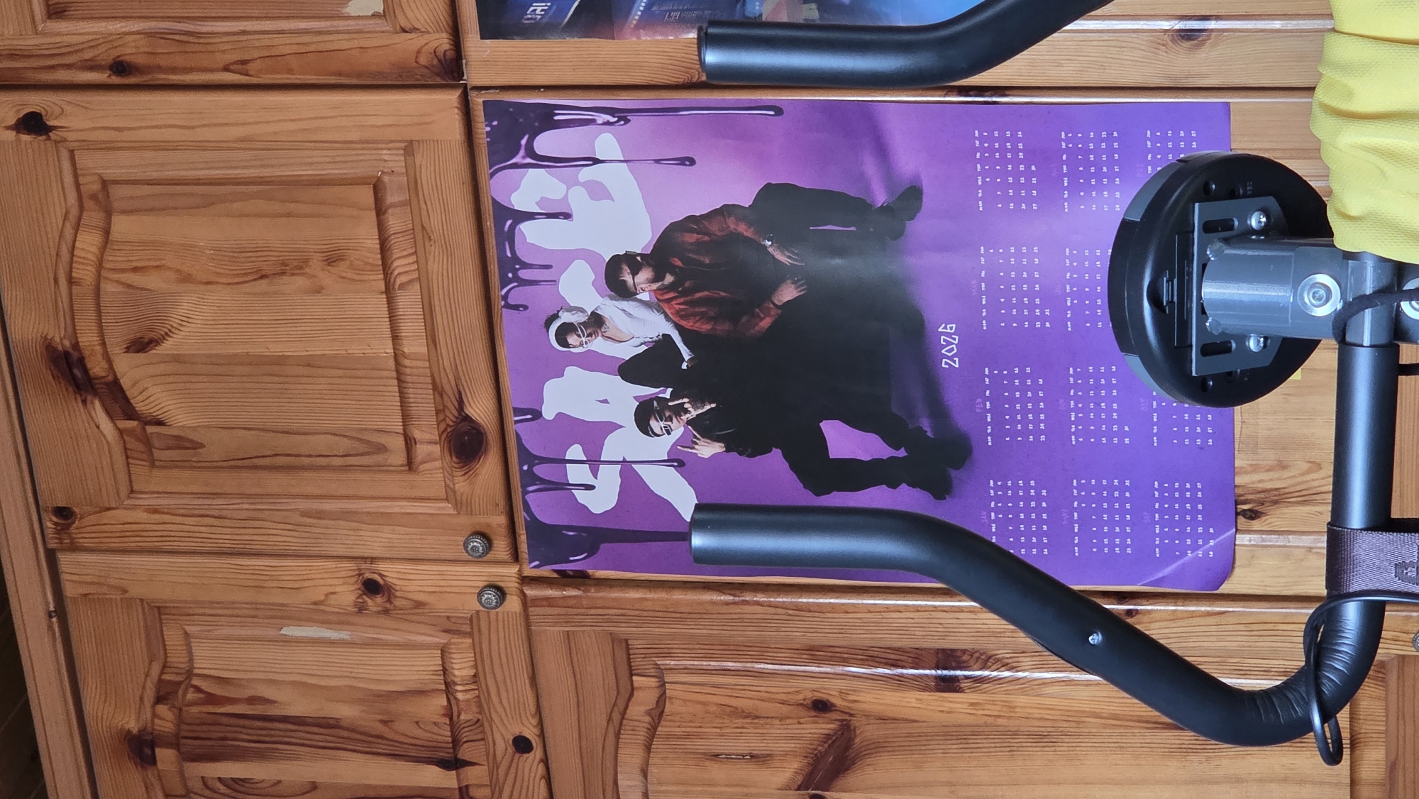 The image displays an indoor setting, likely in Bania, Bulgaria, featuring a section of light brown wooden furniture or wall panels, a purple poster, and parts of an exercise machine. The wooden surface exhibits distinct grain patterns, knots, and a paneled design, along with two small, silver-colored decorative knobs.
Dominating the center is a purple poster, likely a calendar, depicting three stylized figures, appearing to be young men, in various poses. One figure is dressed in a white top with sunglasses, another in a red long-sleeved shirt and glasses, and the third in a black outfit. White, dripping graphic elements are incorporated into the poster's design. The year "2026" is printed vertically on the right side of the poster, accompanied by detailed calendar grids for various months of that year. Visible month abbreviations include JAN, FEB, MAR, APR, MAY, JUN, SEP, OCT, NOV, and DEC, each with days of the week (SUN, MON, TUE, WED, THU, FRI, SAT) and corresponding dates.
In the foreground, obscuring portions of the poster and wood, is part of an exercise machine. A curved black handle or bar extends from the bottom right. Above it, a round, gray and black mechanical component is visible, along with a glimpse of yellow fabric at the top right. A black strap is wrapped around a vertical support of the equipment. The scene is static, with no discernible activity or event taking place, and no specific time of day or emotions are conveyed.