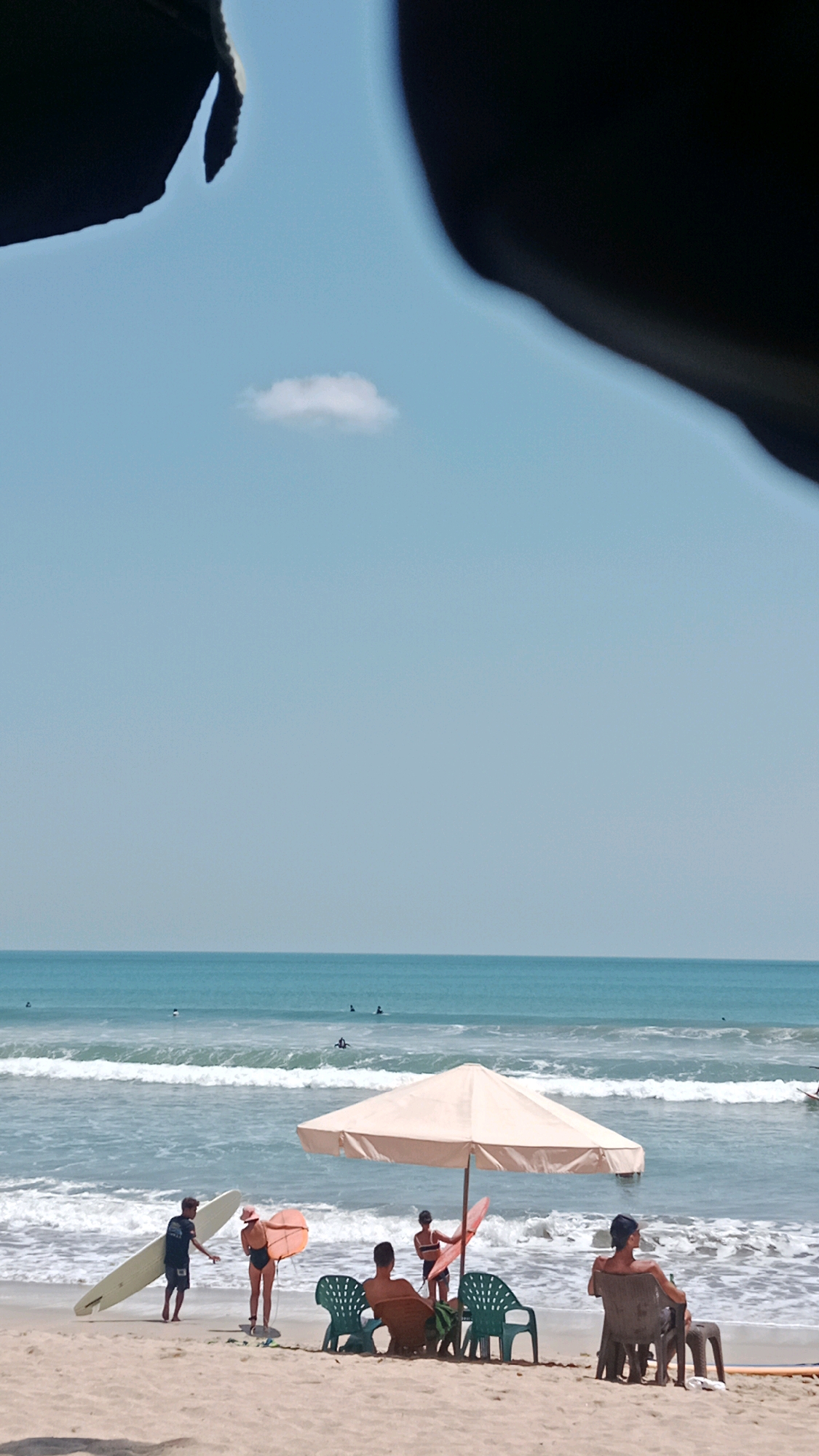 In Kuta, Indonesia, a serene beach scene unfolds under a clear blue sky. The ocean, a vibrant turquoise, meets the sandy shore with gentle waves. A solitary white cloud floats lazily in the vast expanse of the sky. In the foreground, a group of people are enjoying the beach.
Under a large, cream-colored umbrella, three individuals are seated on plastic chairs, facing the ocean. One person, wearing a dark cap, is looking out at the water. Two other individuals, one with short hair and another with a ponytail, are also seated, their gazes directed towards the sea.
To the left, two people are engaged in an activity with surfboards. A man with dark hair and wearing a dark t-shirt and shorts stands with a long, white surfboard. Beside him, a person wearing a pink hat and a black swimsuit holds an orange surfboard. They appear to be interacting with each other, possibly discussing surfing.
In the middle ground, several surfers are visible in the ocean, riding the waves or paddling in the water. The waves are breaking onto the shore, creating a white foam. The overall atmosphere is one of relaxation and enjoyment of a beautiful beach day. The image is framed by dark, indistinct shapes at the top, suggesting the presence of some overhead structure.