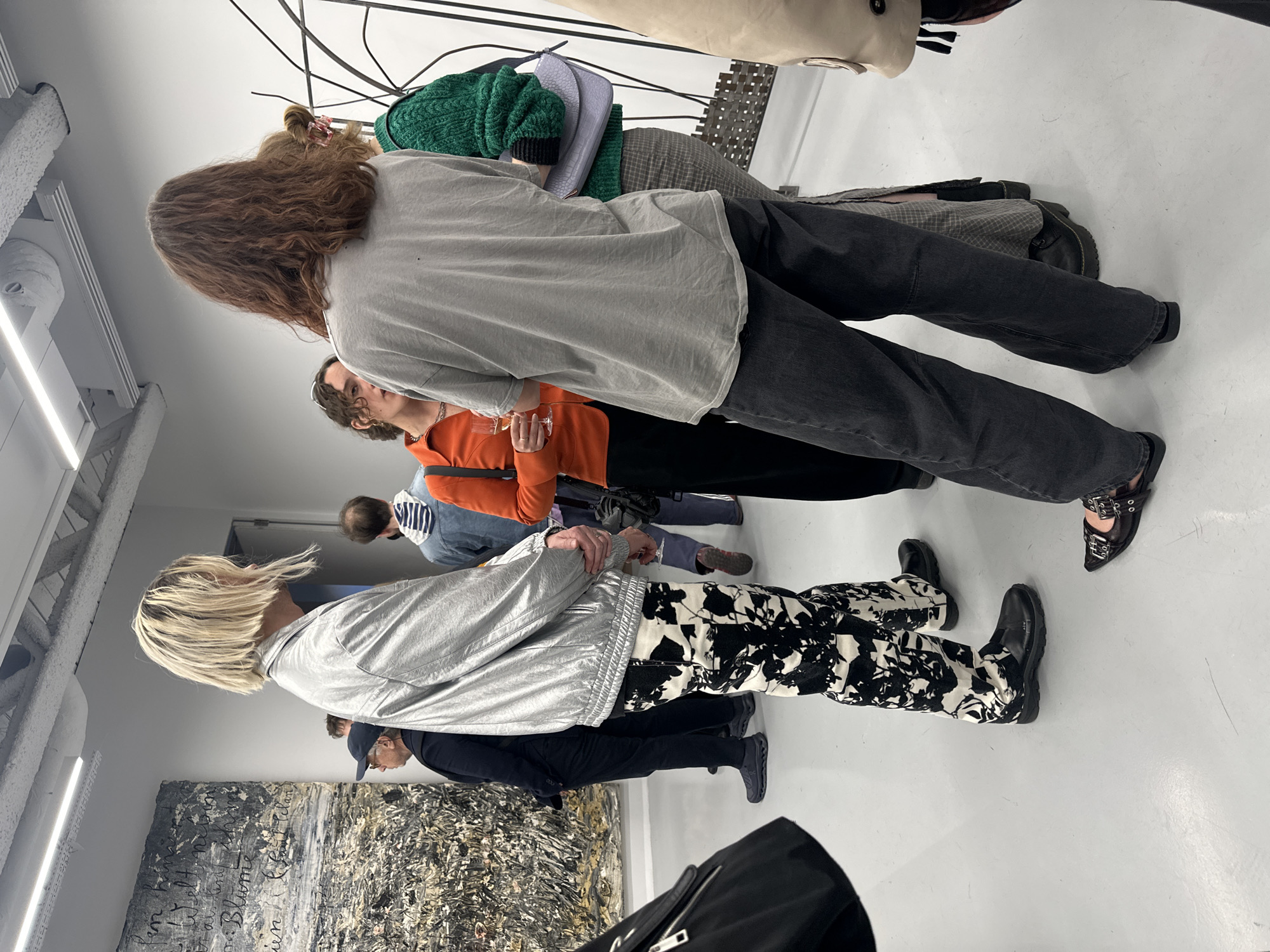 The image, when properly oriented, captures a gathering of people within what appears to be an art gallery or exhibition space in London, United Kingdom. The room features stark white walls, a light grey floor, and an industrial-style ceiling with visible pipes and strip lighting fixtures.
Several individuals are present, observing artworks. In the immediate foreground, facing away, is a person with short, light blonde hair, wearing a metallic silver jacket, black and white abstract-patterned trousers, and black chunky-soled shoes. To their right, seen from behind, is another person with long, wavy brown hair, dressed in a loose light grey t-shirt, dark trousers, and distinctive black shoes with multiple buckles.
Mid-ground figures include a person with curly brown hair wearing a bright orange top, holding a clear glass. Behind them, an individual in a green knitted sweater is partially visible, carrying a light purple bag and wearing plaid trousers and black boots. Other attendees are dispersed throughout the space, including one in a dark jacket and cap, and another in a light blue and white striped top.
On the left wall, a large, dark-toned abstract or mixed-media artwork is displayed, featuring expressive white cursive text. Portions of the text are visible, including what appears to be "Blume" (German for "flower") and other handwritten script, suggesting a European language. Overhead, a complex, wire-like sculpture is suspended. The overall scene suggests an art viewing or exhibition event, with people engaged in contemplating the displays.