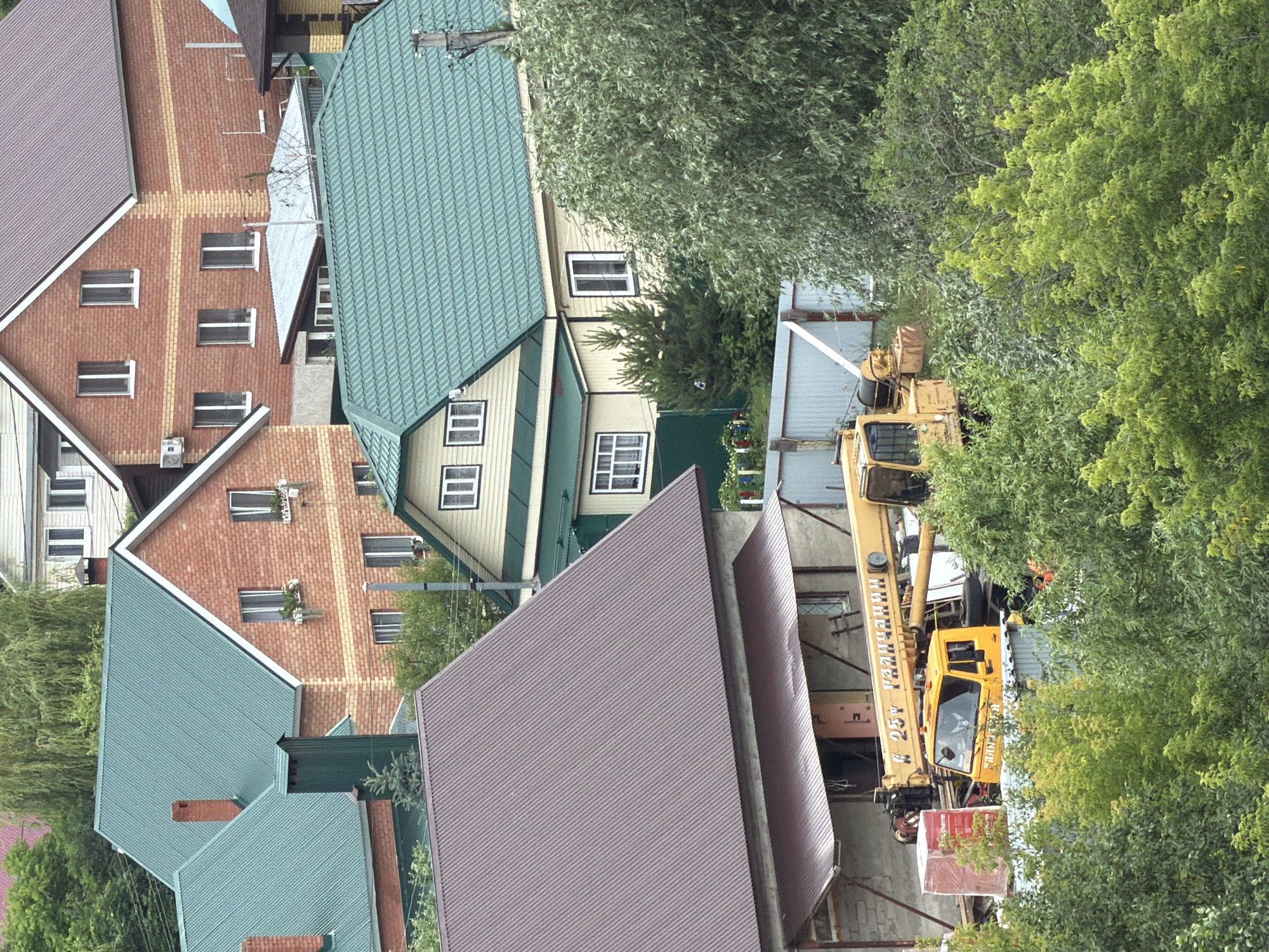 This is a medium-angle, eye-level, full exterior shot of a residential area with a focus on multiple houses and a construction crane. The scene is set in a suburban environment with dense green foliage, including trees and bushes, surrounding the buildings. The houses feature varied roofing materials, with some having green corrugated metal roofs and others with reddish-brown metal roofs. The architecture includes brick and light-colored painted exteriors, with visible windows. In the foreground, a bright yellow crane, identified by Russian text on its boom that reads "К 25т ГАЛЛИАНИНИ" (K 25t GALLIANINI), is positioned over a construction site. The crane's cab is visible, and it appears to be in operation or ready for operation. There are construction materials, including a wrapped red object and what looks like stacks of brick or concrete blocks, near the crane. A smaller, detached building with a brown awning covers an entrance. The overall impression is a construction project underway in a residential neighborhood.