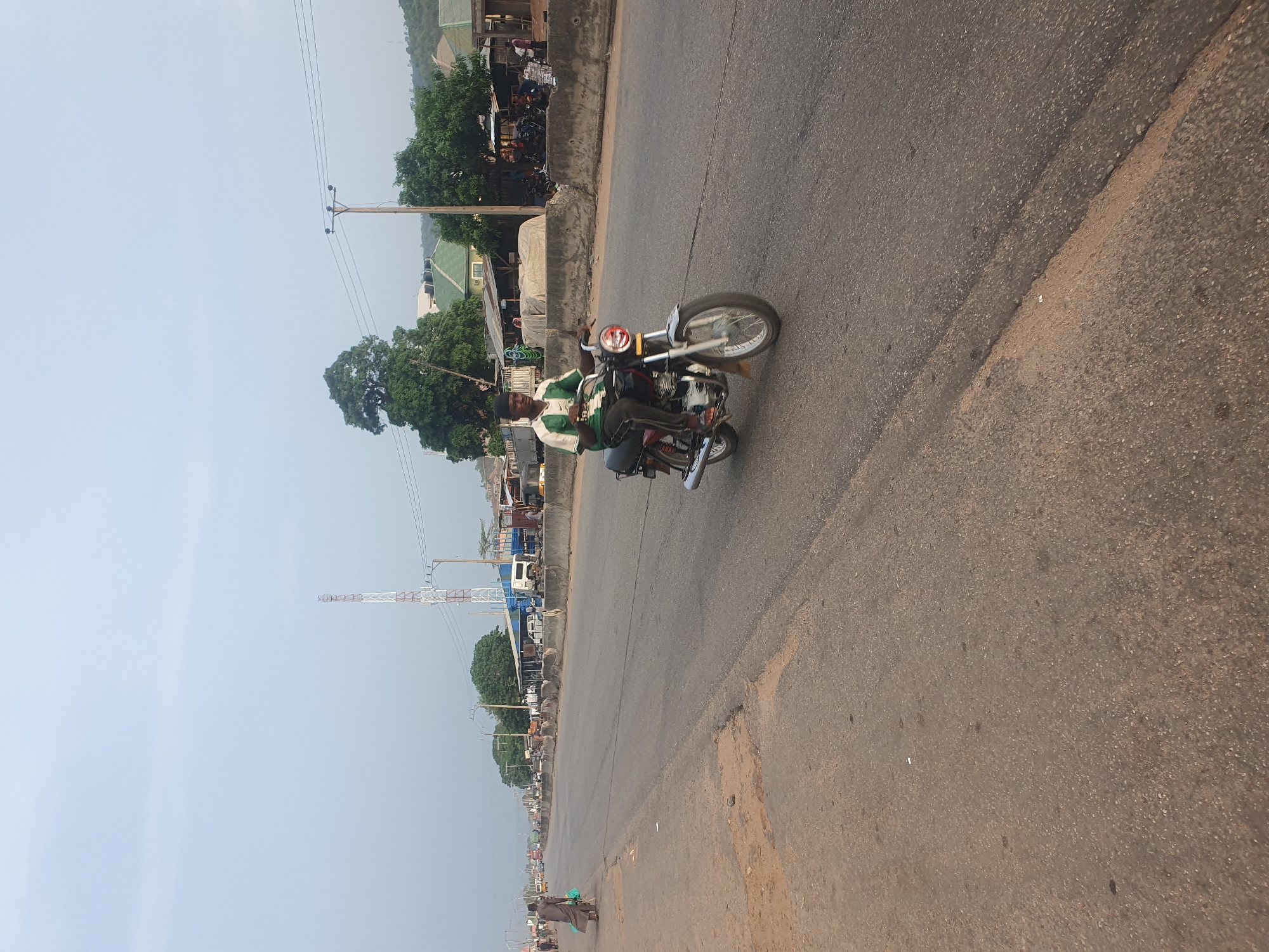 A man is riding a motorcycle on a paved road in Lokoja, Nigeria. The road appears to be a major thoroughfare with traffic in the distance. The man is wearing a green and white jacket and a black hat. Buildings, trees, and a telecommunications tower can be seen in the background.