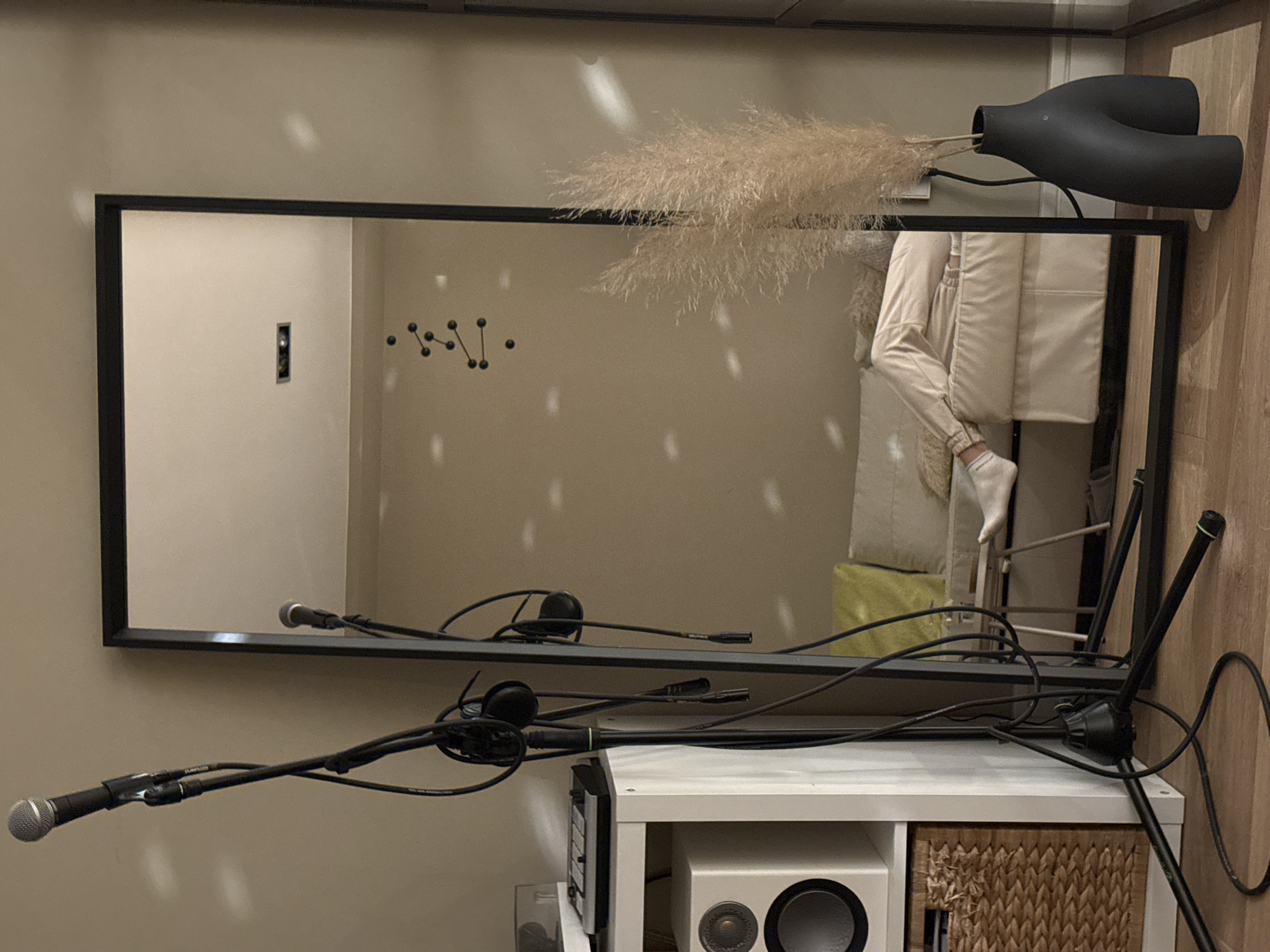 A horizontally oriented image shows a room with a large rectangular mirror in a black frame mounted on a wall. The mirror reflects part of the room, including a person sitting on a chair, visible in the upper right portion of the mirror, and other wall decorations. Above the mirror is a tall black vase holding decorative grass.
In the foreground, in front of the mirror, is a white shelving unit with a speaker and a wicker basket inside. Black microphones and cables are positioned in front of the mirror and shelving unit, obscuring parts of the view. The overall color scheme is neutral with beige walls and furniture. The specific location in Barcelona, Spain, is not identifiable from the image.