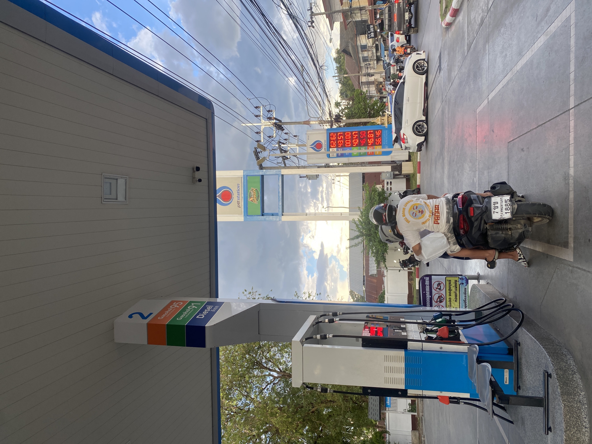 Here is a description of the image:
The image captures a scene at a PTT Station, a petrol station in Baan Mae Nam, Thailand. A person on a motorcycle is parked at pump number 2, presumably refueling. The pump displays options for Gasohol 95, Gasohol 91, and Diesel. In the background, a PTT station sign with an Amazon logo is visible. The gas price board displays the gas prices on offer.
Vehicular traffic, including a white car, lines the road. Power lines intersect the sky above the station, and buildings line the back of the street with trees.