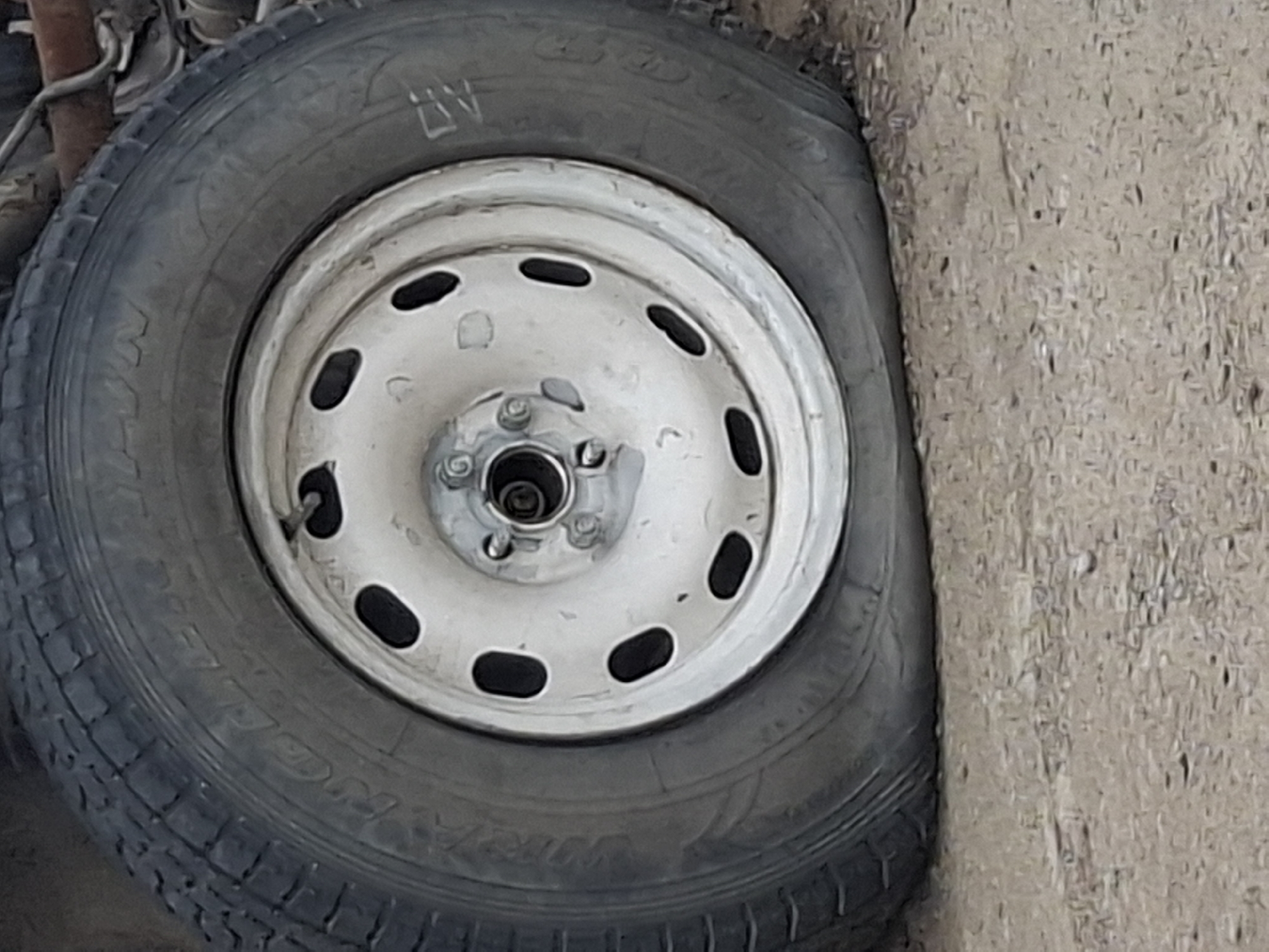 An overhead shot captures a deflated tire on a vehicle, suggesting a flat tire incident. The tire features a silver rim with several oval-shaped cutouts. The tire itself is black and appears worn. The background is out of focus, but the ground beneath the tire is visible and appears to be a rough, unpaved surface. There are no people or other objects clearly identifiable in the image. No specific location in Almaty, Kazakhstan, can be identified from the visual information provided.