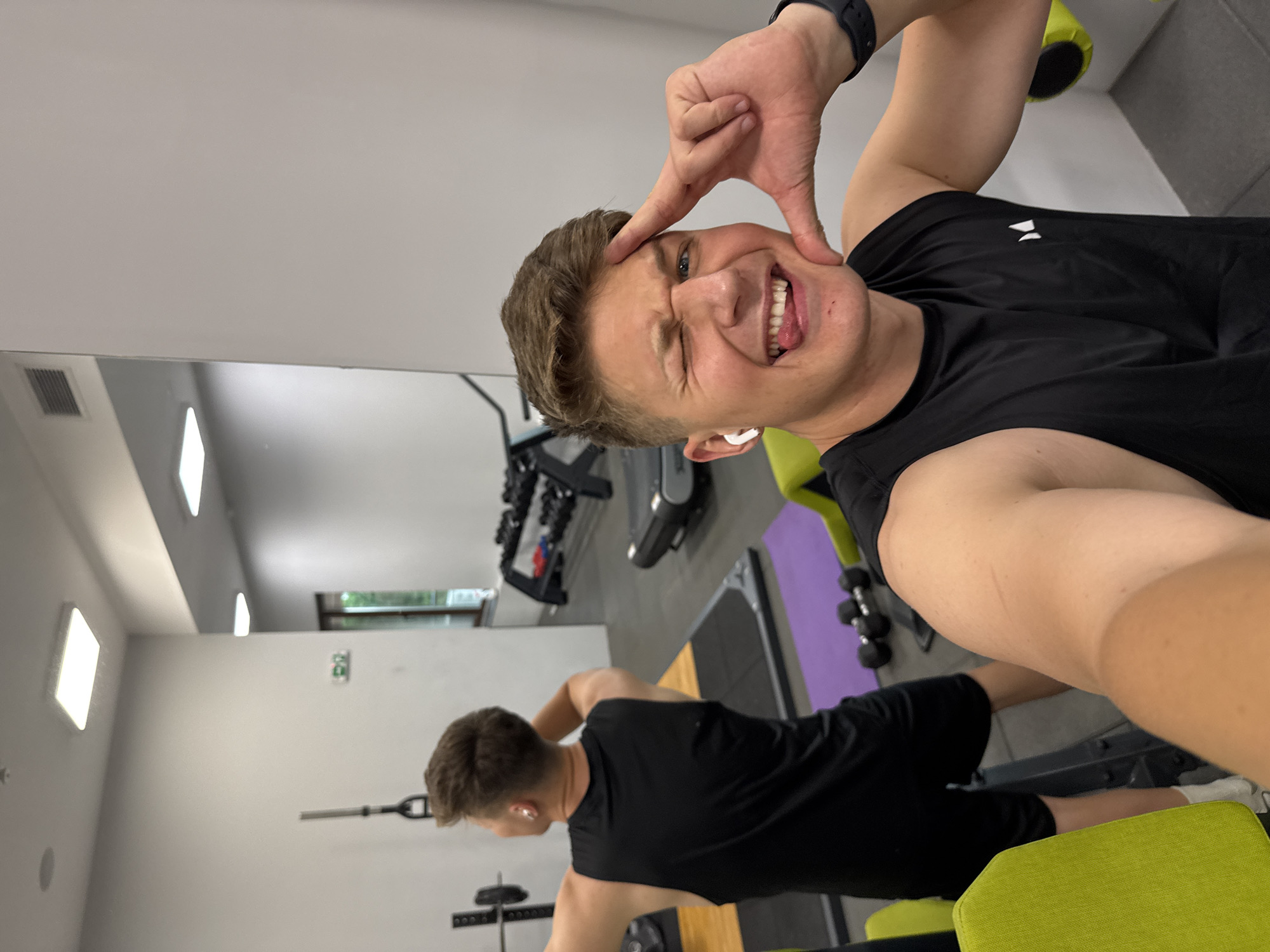 A selfie-style image captures a man in a gym. He's dressed in a black tank top and wears a sports watch on his left wrist and a white earbud in his left ear. He is making a playful face at the camera with a wide smile, sticking out his tongue, winking, and framing his face with his hand. The gym has exercise equipment, a mirror reflecting part of the room, and what appears to be another person in the background. The location cannot be inferred from the image provided.