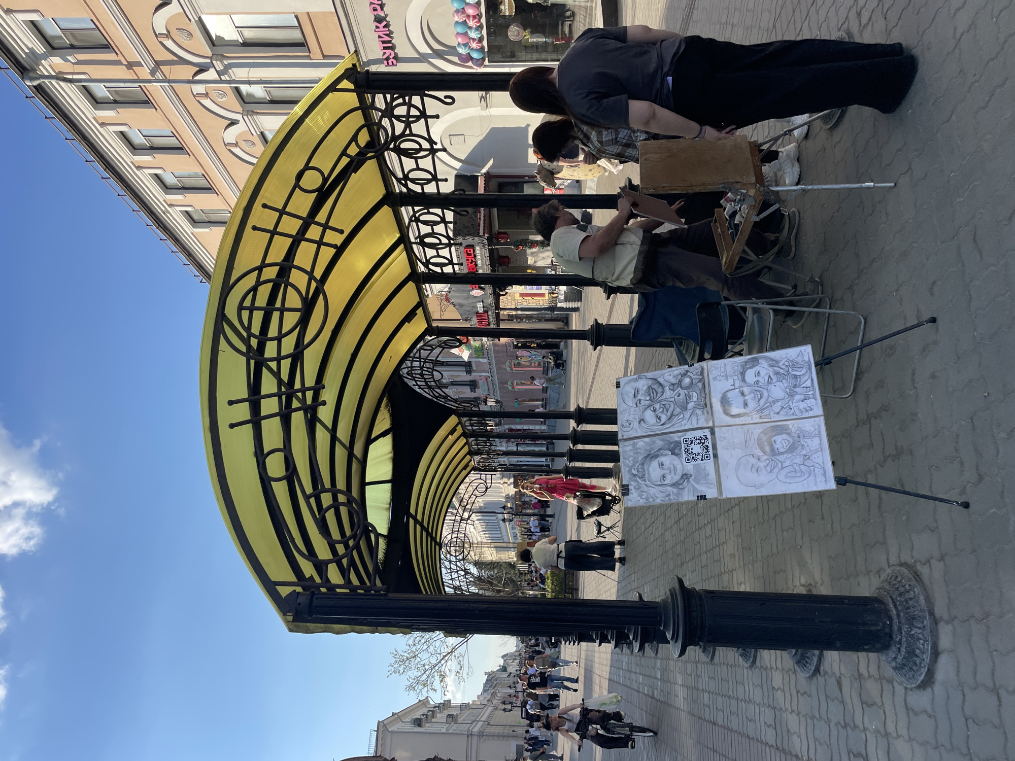 The image captures a street scene in Kazan, Russia. A yellow awning with black metal framework provides cover to a section of the paved pedestrian walkway. Underneath the awning, an artist sits on a chair, drawing a portrait of two women who are observing him. His artwork is displayed on an easel nearby. People stroll along the walkway in both directions. Buildings line either side of the street. The sky is clear and blue.