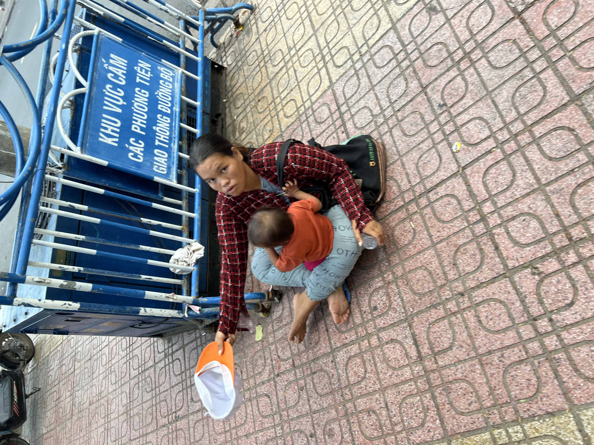 A woman and a baby are sitting on the sidewalk in front of a temporary barrier made of metal poles and a blue panel. The woman is wearing a red and black plaid shirt and grey pants. She is holding an orange and white hat in her left hand and a baby is sitting on her lap. The baby is wearing an orange shirt. The sidewalk is paved with red tiles. Behind the barrier, there is a generator. A sign on the barrier reads in Vietnamese: " khu vực cấm các phương tiện giao thông đường bộ" which translates to "Forbidden area for vehicles". The scene appears to be in a public area in Nha Trang, Vietnam, likely near a construction site or an event.