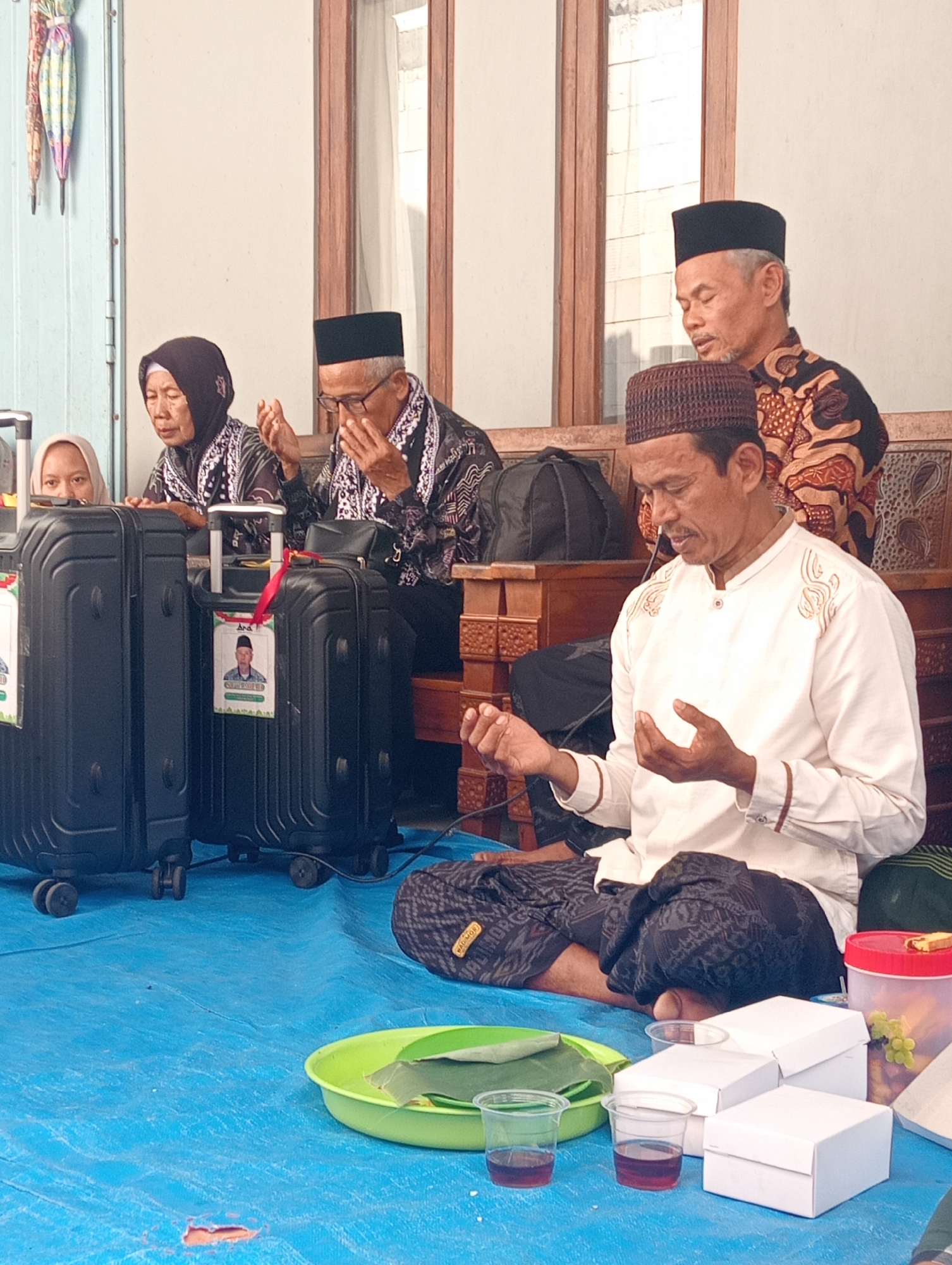 A group of people are gathered on a blue tarp, seemingly engaged in a prayer or ritual. In the foreground, a man in a white shirt and patterned sarong sits with his hands cupped, eyes closed in prayer. To his left, a woman in a black hijab and patterned top also has her hands raised, her eyes closed. Behind them, another man, also in traditional attire, is visible, his gaze downcast. Two large black suitcases are positioned to the left of the seated figures, one with a photograph of a man attached. In front of the prayer group are several white boxes, two plastic cups containing a dark liquid, and a green bowl holding banana leaves. The background suggests an indoor or semi-outdoor setting with wooden architectural elements and light filtering through windows. The scene appears to be in Boyolali, Indonesia, given the context provided.