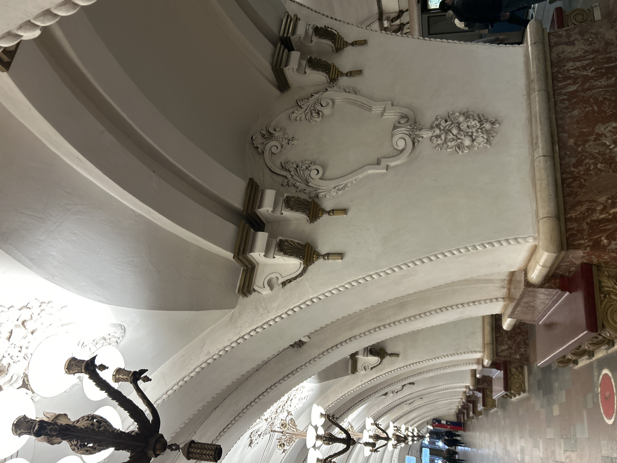 The image shows the interior of a metro station in an unknown city. The architecture features ornate white arches decorated with gilded details. The perspective is from a low angle, looking up the length of the station. Chandeliers hang from the center of each arch. The station is populated with people walking in the distance. The floors are made of a mixture of marble and tile.