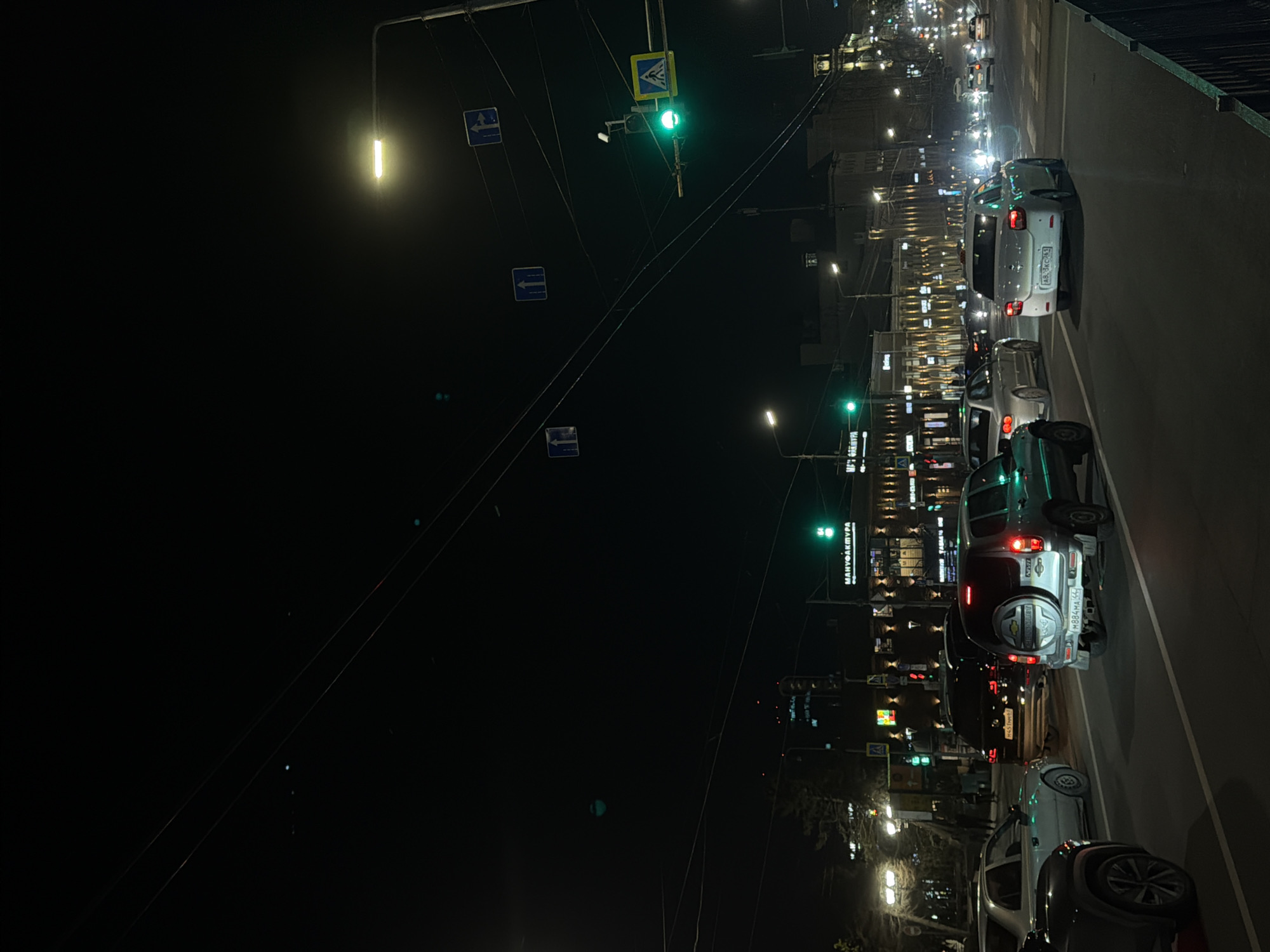 At night, a street in Rostov-on-Don, Russia, is lined with parked cars. Streetlights illuminate the road, and buildings with lit windows stand in the background. Traffic lights show green for pedestrians. Overhead wires extend across the scene.
