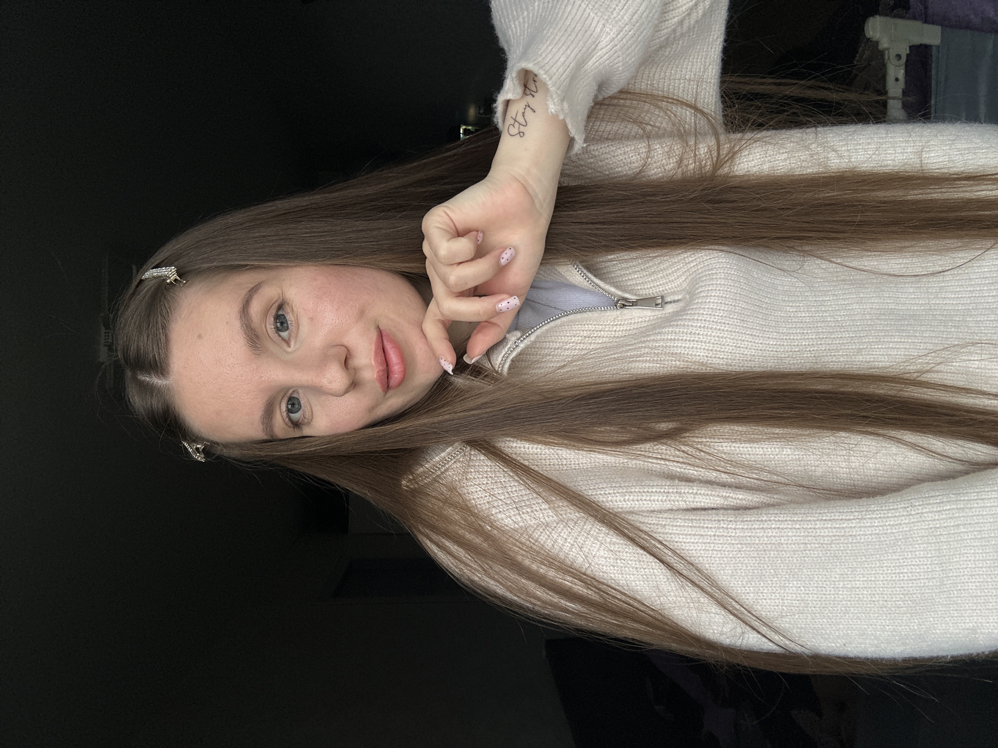 A fair-skinned woman with long brown hair is positioned against a dark background. She wears a light-colored knit sweater with a zip-up collar. A hair clip is visible in her hair near her forehead. Her right hand is raised, with a tattoo visible on her wrist. Her nails are painted a light color with small dark dots. The image does not contain any visual information that confirms it was taken in Klin, Russia.