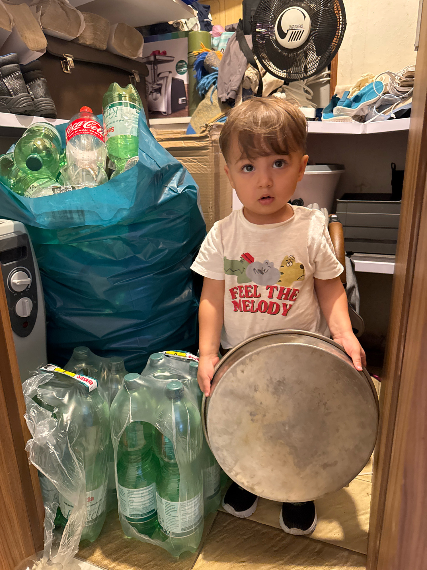 A young child with brown hair and brown eyes is standing in a cluttered closet in Überherrn, Germany. The child is wearing a white t-shirt with cartoon animals and the words "FEEL THE MELODY" printed on it, and black and white sneakers. They are holding a large, round, metal baking pan. To the child's left, a large blue recycling bag is filled with plastic bottles, including Coca-Cola bottles and green bottles of Saskia mineral water. To the child's right, there are stacks of plastic-wrapped green bottles of Saskia mineral water. In the background, shelves are visible, holding various items including boots, a fan, and other miscellaneous objects. A gray electric heater is positioned next to the blue recycling bag. The closet appears to be a storage area with a variety of household items.