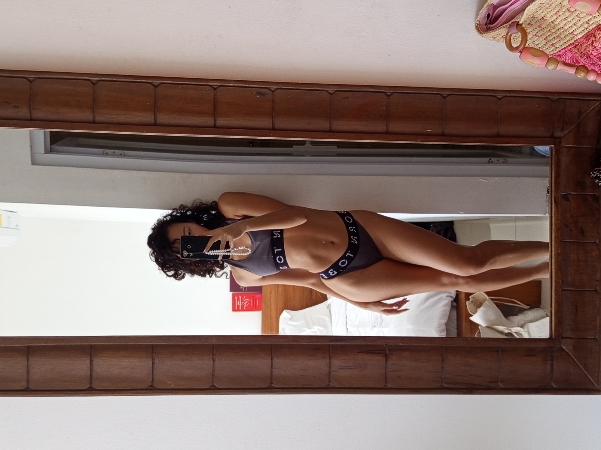 A person with curly black hair is visible in a mirror. The person is holding a cell phone and wearing a gray sports bra and matching underwear with writing on the bands. The mirror is surrounded by a wooden frame with rectangular sections. The background shows a white wall, a bed with white sheets, and other objects on a table next to the bed. There is a straw bag with pink accents in the upper right corner. The location and city are unknown.