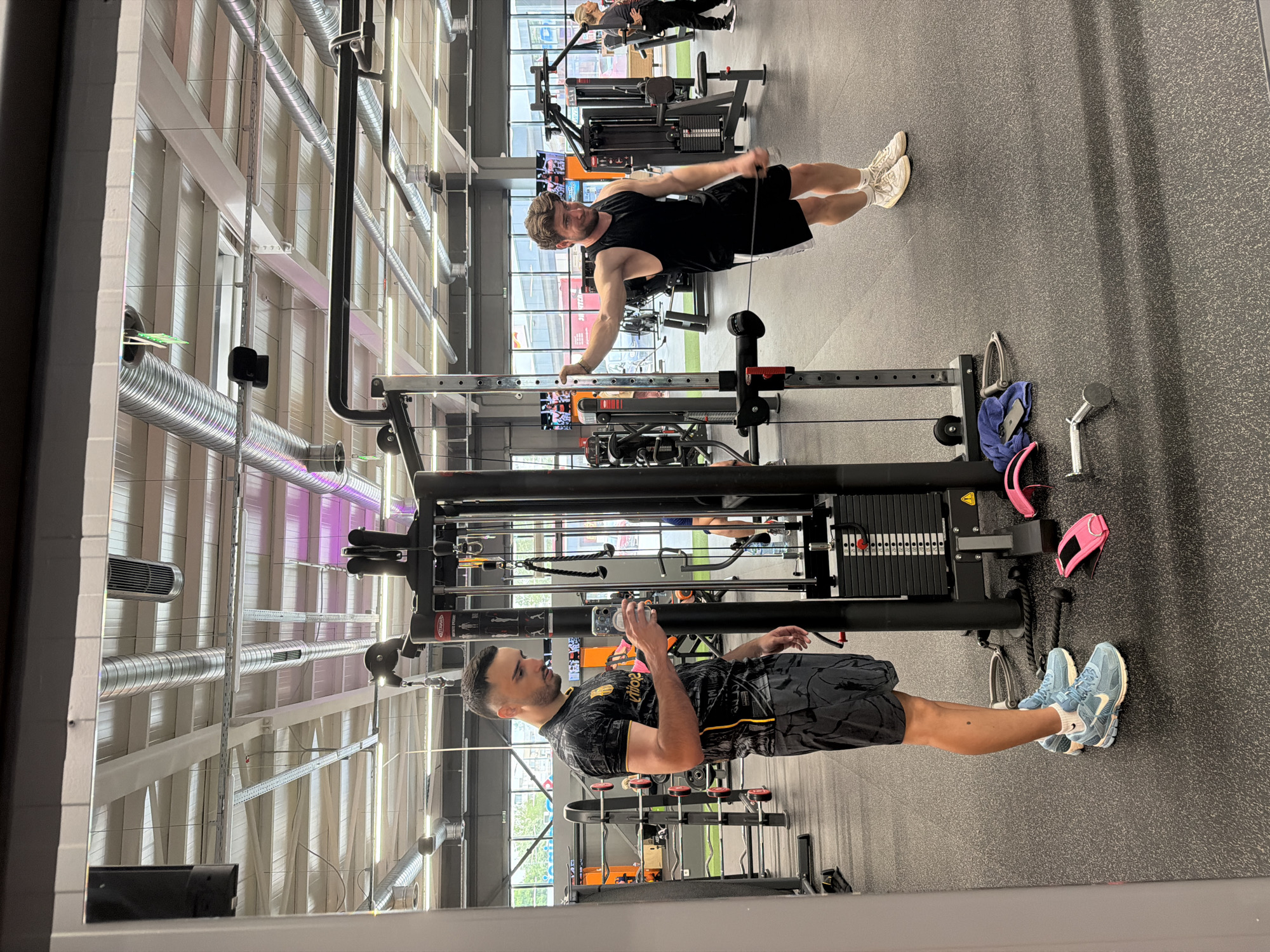 A medium indoor shot depicts a gym scene with two men and several pieces of exercise equipment. The gym has a high ceiling with exposed silver ductwork and white panels, indicating a modern industrial style.
In the foreground, a man in a black and gray patterned t-shirt and black shorts stands near a cable machine. He is holding a smartphone, potentially taking a picture or video. He is wearing light blue sneakers with white socks.
In the background, another man wearing a black tank top and shorts is performing a cable row exercise. He is pulling the cable towards his chest while standing with one hand on a horizontal bar. The gym includes weight machines, dumbbells, towels, and personal items like phones and gloves scattered around.
The setting appears to be a fitness center in Plovdiv, Bulgaria, characterized by its modern equipment and layout, common in contemporary gyms. The floor is a speckled dark gray or black material.