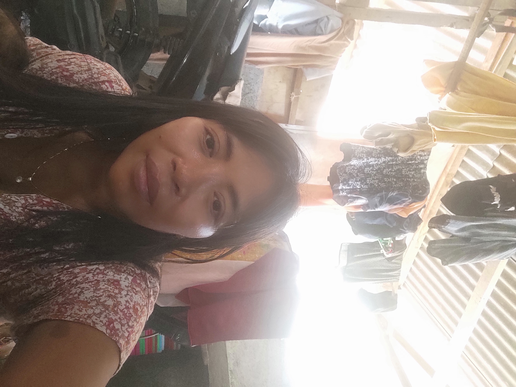A selfie shows a woman with long, dark hair and light brown skin. She is wearing a light-colored dress with a floral pattern. The woman is looking at the camera and smiling. In the background, there are clothes hanging on a clothesline, and a motorcycle is partially visible. The scene appears to be outdoors, possibly in a residential area in the city of Sakra, Indonesia. The lighting suggests it is daytime, with bright sunlight coming from the right side of the image.