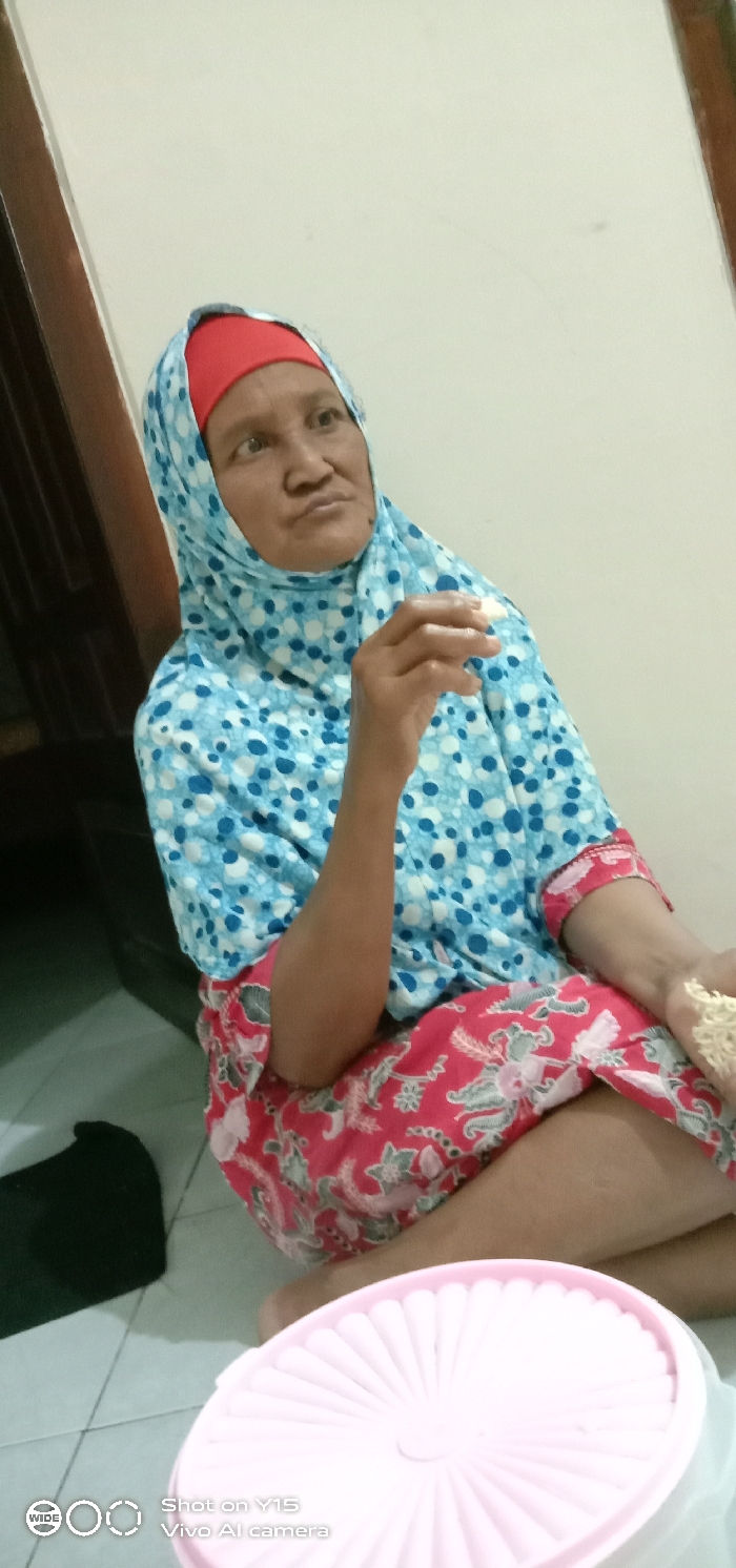 An adult female is depicted seated on a light-colored tiled floor within an indoor environment in Ranugedang, Indonesia. She wears a light blue and white polka-dotted hijab over a red undercap, a matching long-sleeved top, and red patterned lower garments. Her right hand is raised, holding a small, irregularly shaped, light-colored food item, possibly a cracker or fried snack. Her left hand rests on her lap, containing more of the same food. Her gaze is directed towards the upper right of the frame. The background consists of a plain, light-colored wall with a dark vertical doorframe visible on the left. In the lower foreground, a pink round container with a ribbed lid is present on the floor, adjacent to an indistinct dark object.