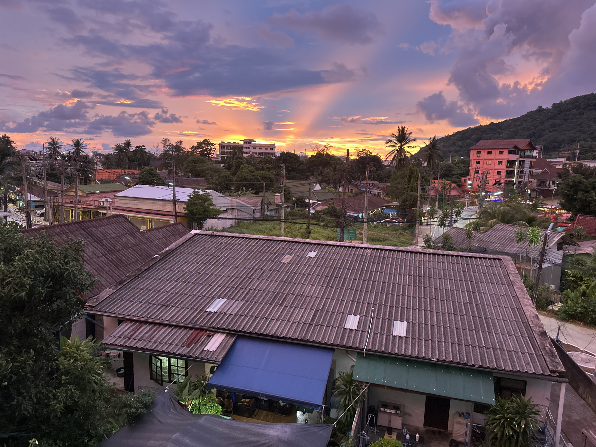 The image depicts a panoramic view of Karon, Thailand, during a vibrant sunset. The sky is a canvas of dramatic colors, with hues of pink, orange, and purple clouds illuminated by the setting sun's rays. In the foreground, a cluster of houses with tiled roofs dominates the scene, their muted tones contrasting with the colorful sky. Several houses are visible, including one with a prominent blue awning and another with a green awning. Beyond the immediate rooftops, the landscape unfolds into a dense tapestry of trees and palm trees, characteristic of a tropical environment. Scattered throughout the scene are various buildings, including a few taller, multi-story structures that suggest a developing urban area. A lush green hillside rises in the background, completing the picturesque backdrop. The overall impression is one of a serene, tropical evening in a coastal town.