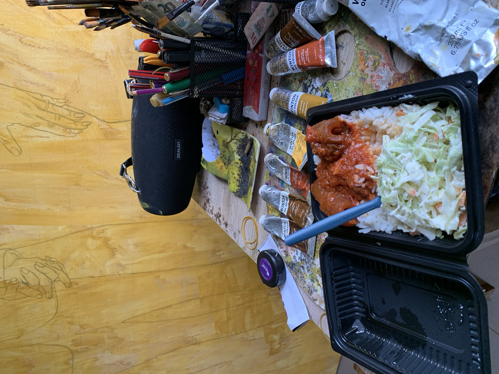 The image depicts a close-up view of a desk cluttered with art supplies and a meal. The background features a yellow wall with faint charcoal sketches of hands. On the desk, a black portable speaker with the "ZEALOT" logo is visible, along with various paint tubes, brushes in a holder, and pens. In the foreground, a black plastic takeout container holds a meal consisting of rice topped with a red sauce and meat, and a side of coleslaw. A blue utensil is placed in the coleslaw. The overall impression is of an artist's workspace where a meal has been placed. The location is not specified in the image or the provided context.