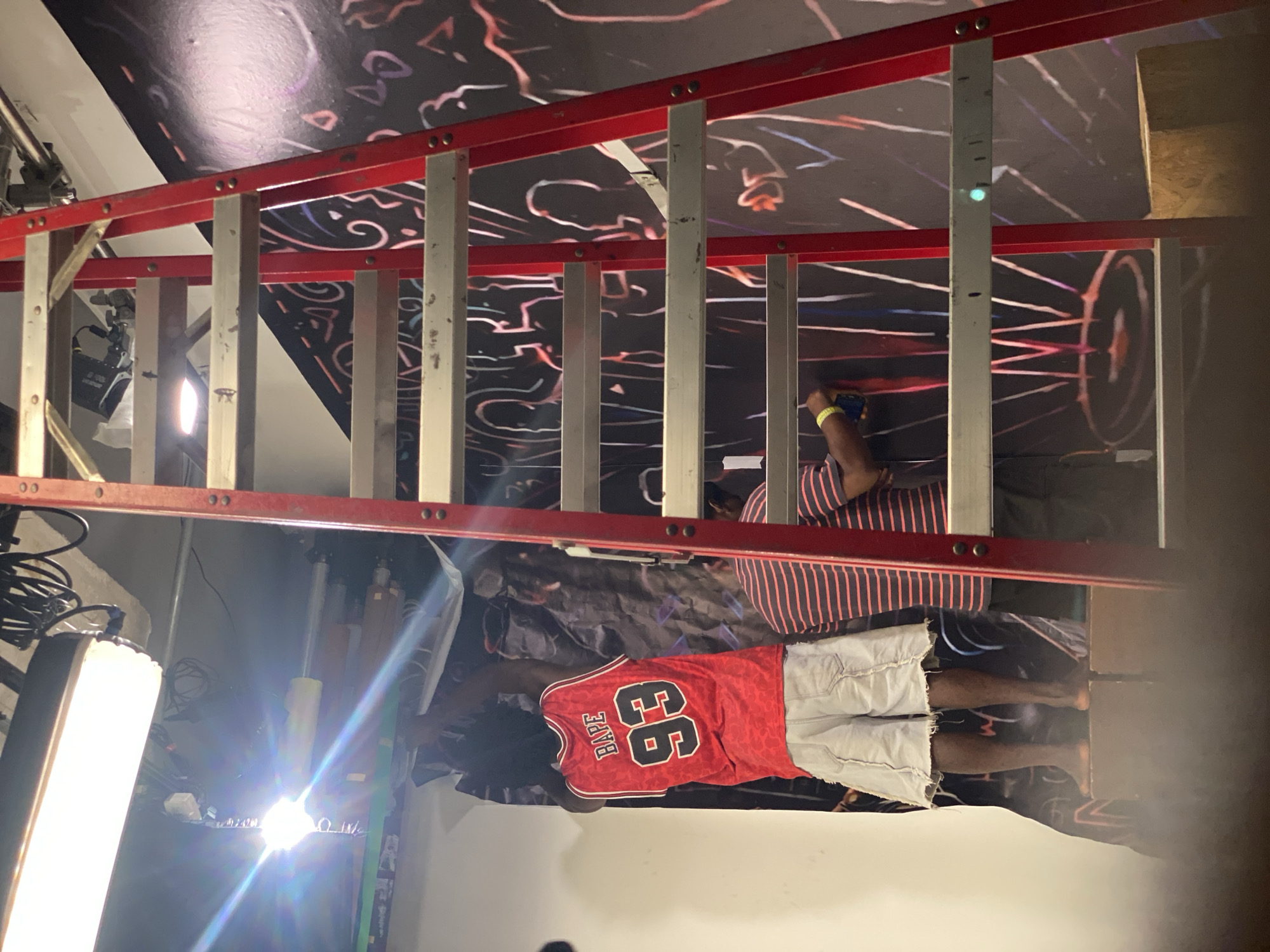 The image captures an indoor scene, likely a studio or creative space, where two individuals are engaged in a project involving a large, dark backdrop adorned with vibrant, abstract, neon-like designs.
A prominent red and silver extension ladder diagonally crosses the frame. On the lower half of the image, a person is seen from behind, wearing a red basketball jersey with "BAPE 93" printed in white on the back, along with white shorts. They are barefoot and appear to be standing on a lower platform or the floor, with their left arm raised as if painting or adjusting something on the backdrop.
Another person is partially visible behind the ladder on the right side, wearing a striped shirt. This individual is positioned higher, likely on the ladder, holding a dark-colored smartphone in their left hand and wearing a yellow wristband. Their right hand is also extended towards the backdrop, indicating participation in the activity, possibly fine-tuning the design or taking reference images.
The backdrop itself features swirling, linear, and geometric patterns in various bright colors including white, red, blue, and purple, creating a dynamic visual effect. Studio lighting equipment, including a softbox on the left and a spotlight casting bright flares, illuminates the scene, suggesting a professional setup for photography, videography, or art creation. Cables are also visible.
The scene conveys a sense of active artistic endeavor in a controlled, artificially lit environment. The location is indicated to be Ikeja, Nigeria.