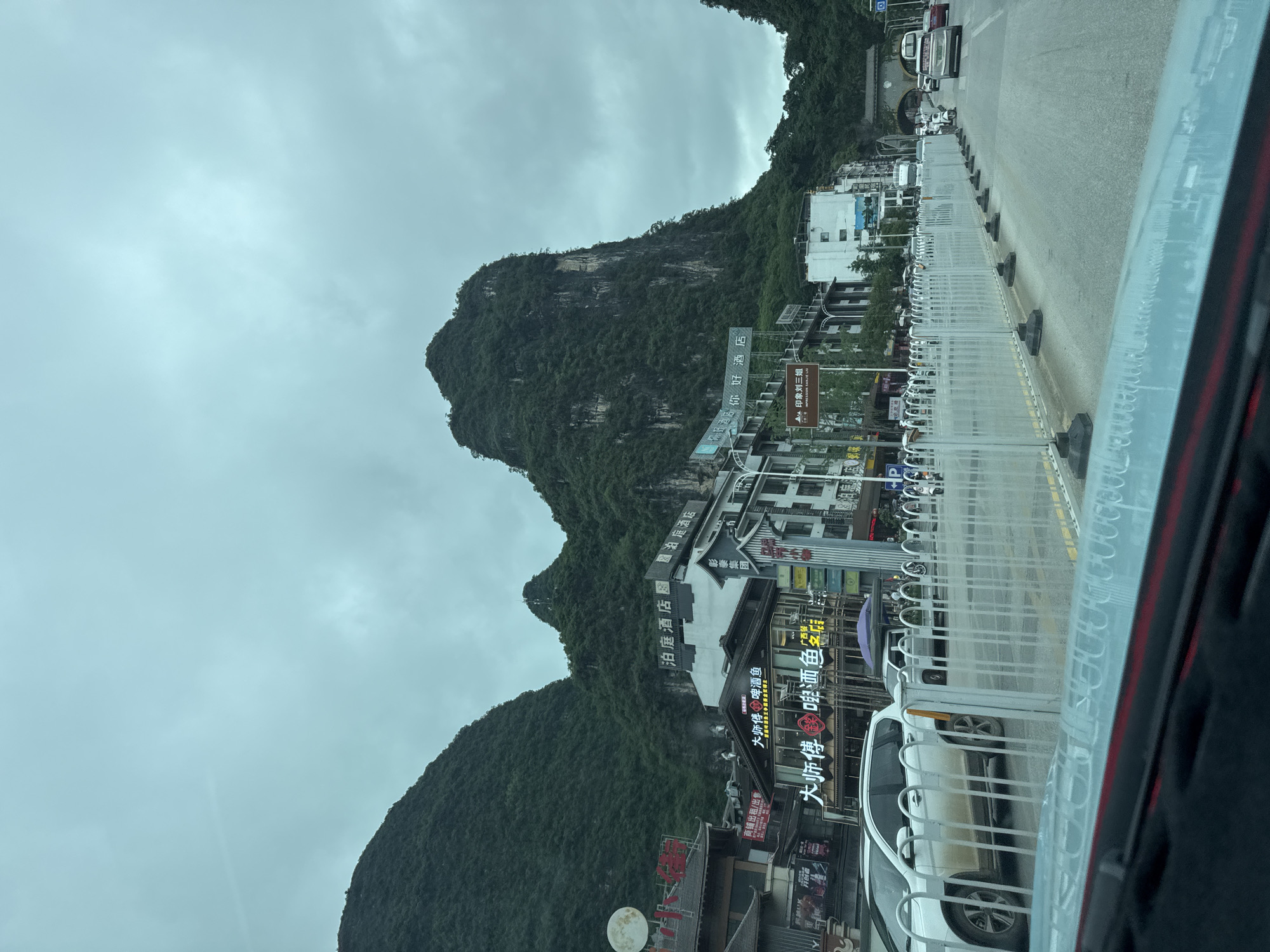 The image shows a view of a street in Guilin, China, with a prominent mountain in the background. The weather appears overcast. A road is visible with a white metal fence running along it. Various buildings line the street, with signs in Chinese characters and advertisements for businesses, including restaurants and hotels. A white car is parked on the side of the street. A sign visible mentions "Impression Sanjie Liu," a well-known outdoor performance show near Guilin.