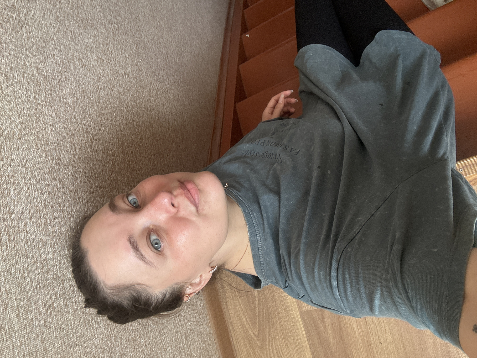 This is a medium shot of a young woman with light skin and blue eyes, lying on her side on a carpeted floor. She is wearing a grey t-shirt with faded text that appears to read "Vintage style FASHION". The woman is looking directly at the camera with a neutral expression. She has dark brown hair that is pulled back from her face, and she is wearing hoop earrings and a delicate necklace. The background shows wooden stairs and a carpeted floor. The lighting suggests it is daytime, with natural light illuminating the scene. The overall impression is casual and intimate, as if the photo was taken spontaneously.
