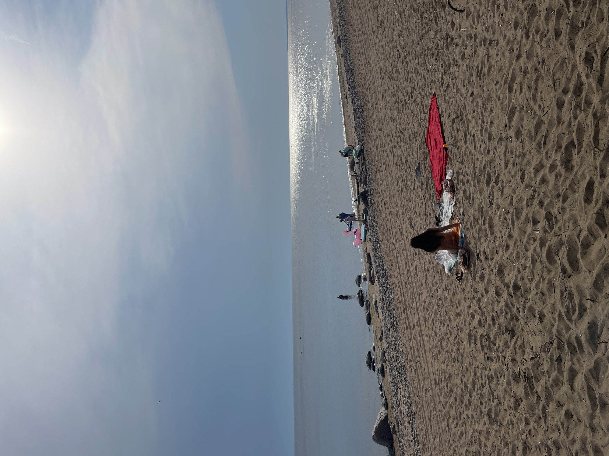A wide shot captures a beach scene under a bright, hazy sky. In the foreground, a person sits on a towel on the sand, facing away from the camera, dressed in a swimsuit. To their right, a red towel is spread out on the sand. The beach transitions to a rocky shoreline bordering a calm body of water. Several people are visible in the water, some wading and others standing. In the distance, the sun reflects brightly off the water's surface. Several inflatable objects, including a pink flamingo, are present near the water's edge. The location is identified as the city of Luzhki, Russia.