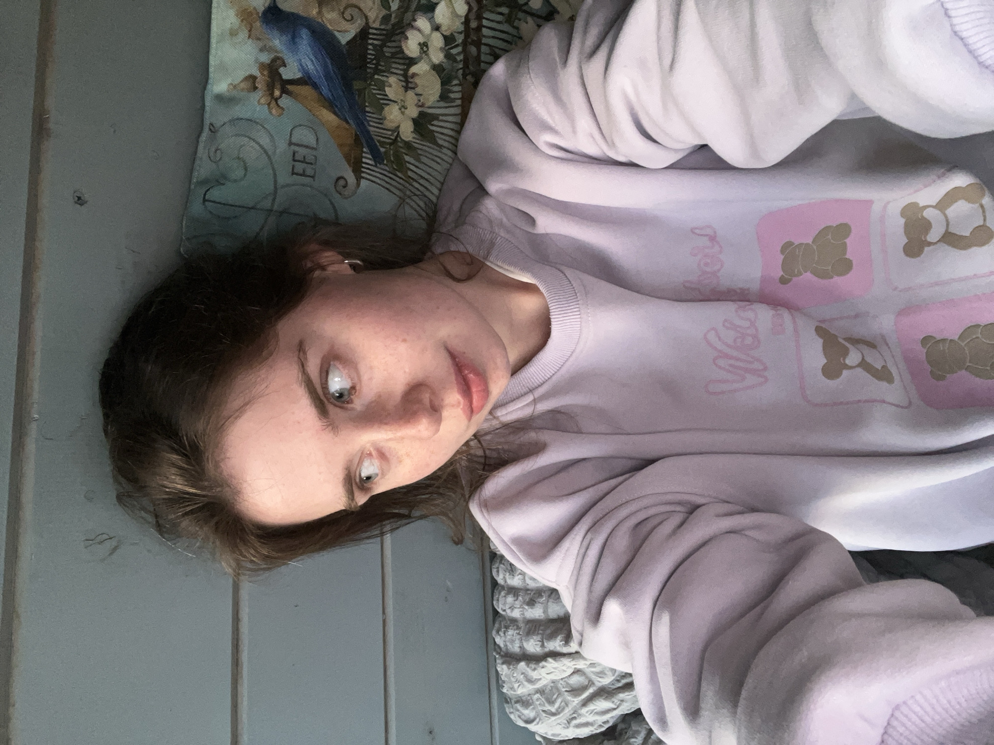 A young woman lies on her side, facing the camera. She has fair skin, brown hair, and freckles. She is wearing a light purple sweatshirt with teddy bear designs on the front. She is resting on a pillow with a blue bird design and floral patterns. Next to her is a light blue wall with vertical panels. The specific location within the city of Chaikovsky, Russia, cannot be determined from the image.